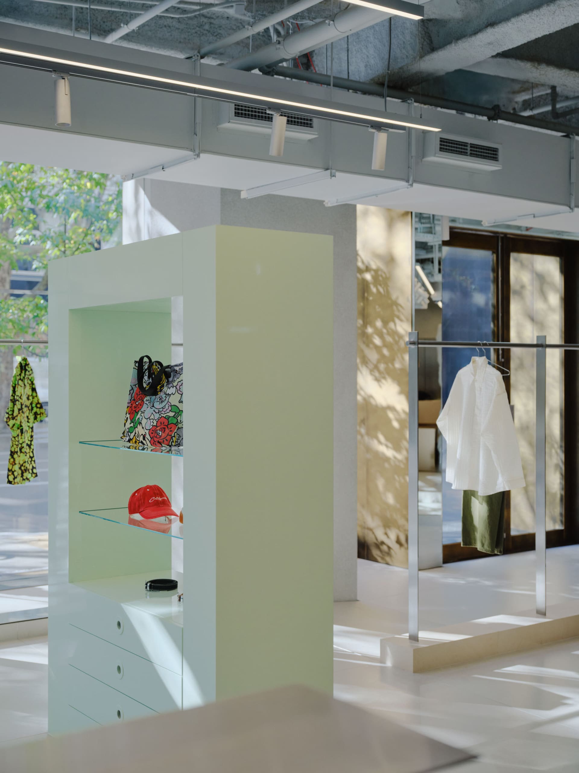 A modern clothing retail store displays clothing on glass shelves, with concrete columns and a metallic rail.