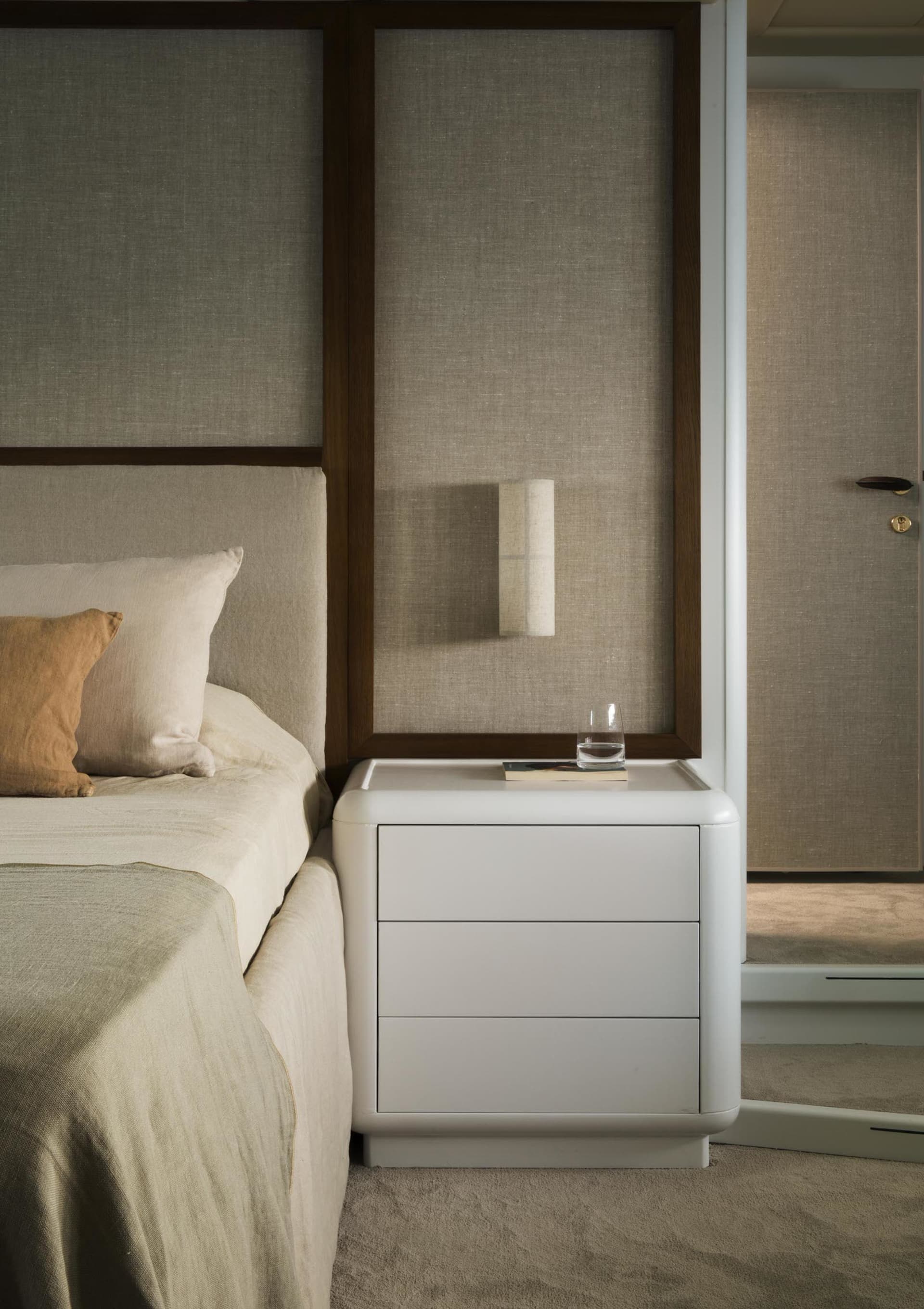 A minimalist bedroom with a white bed and nightstand, featuring a glass and metal lamp and a book.