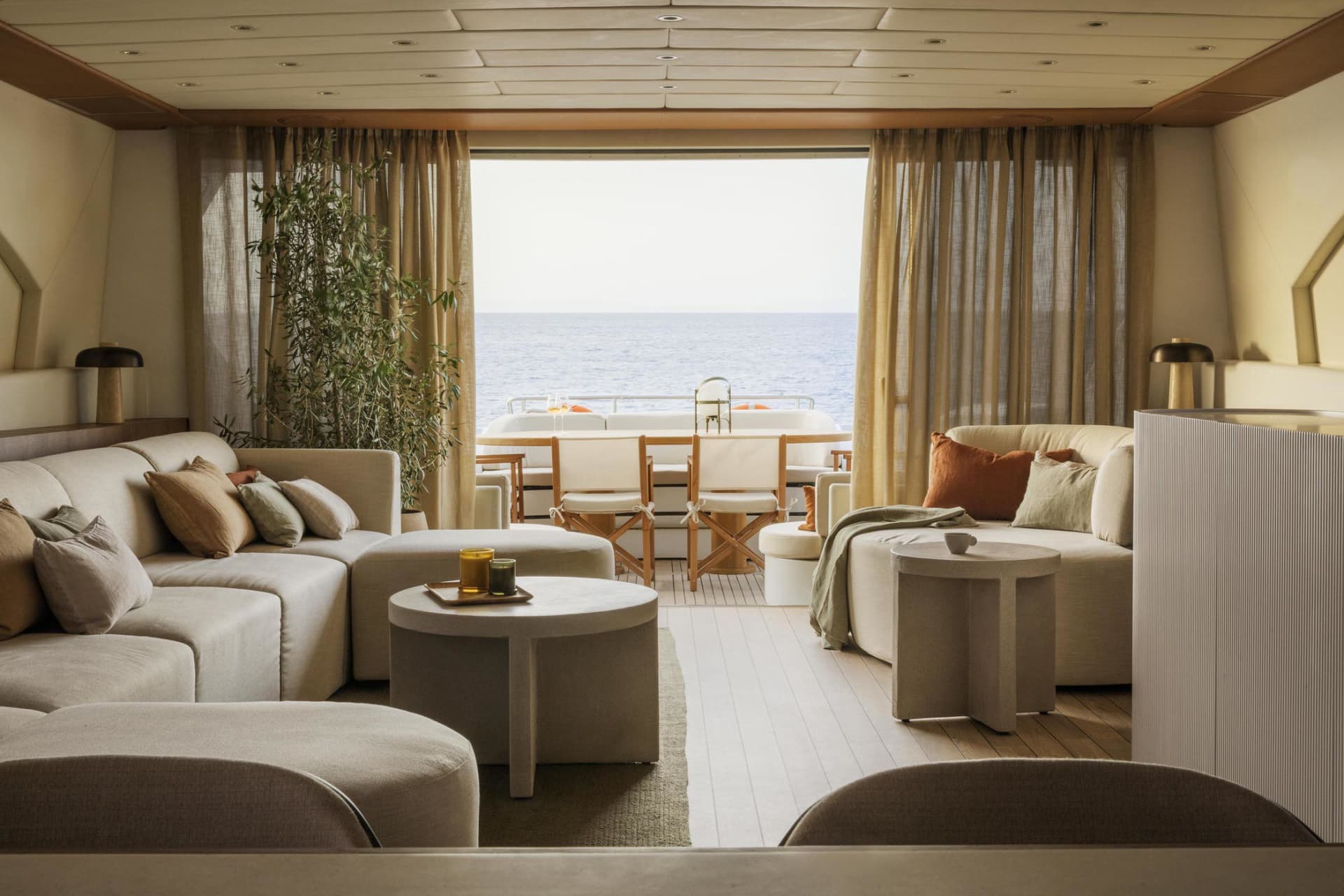 A stylish, modern living room with neutral-toned sofas, ottomans, and tables, featuring large windows with curtains overlooki