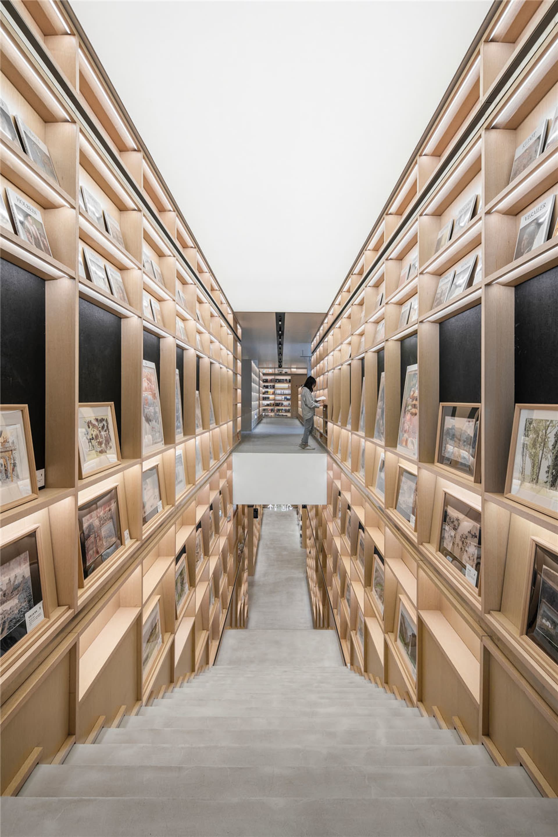 A modern library with wooden shelves filled with books, viewed from the staircase leading upwards.