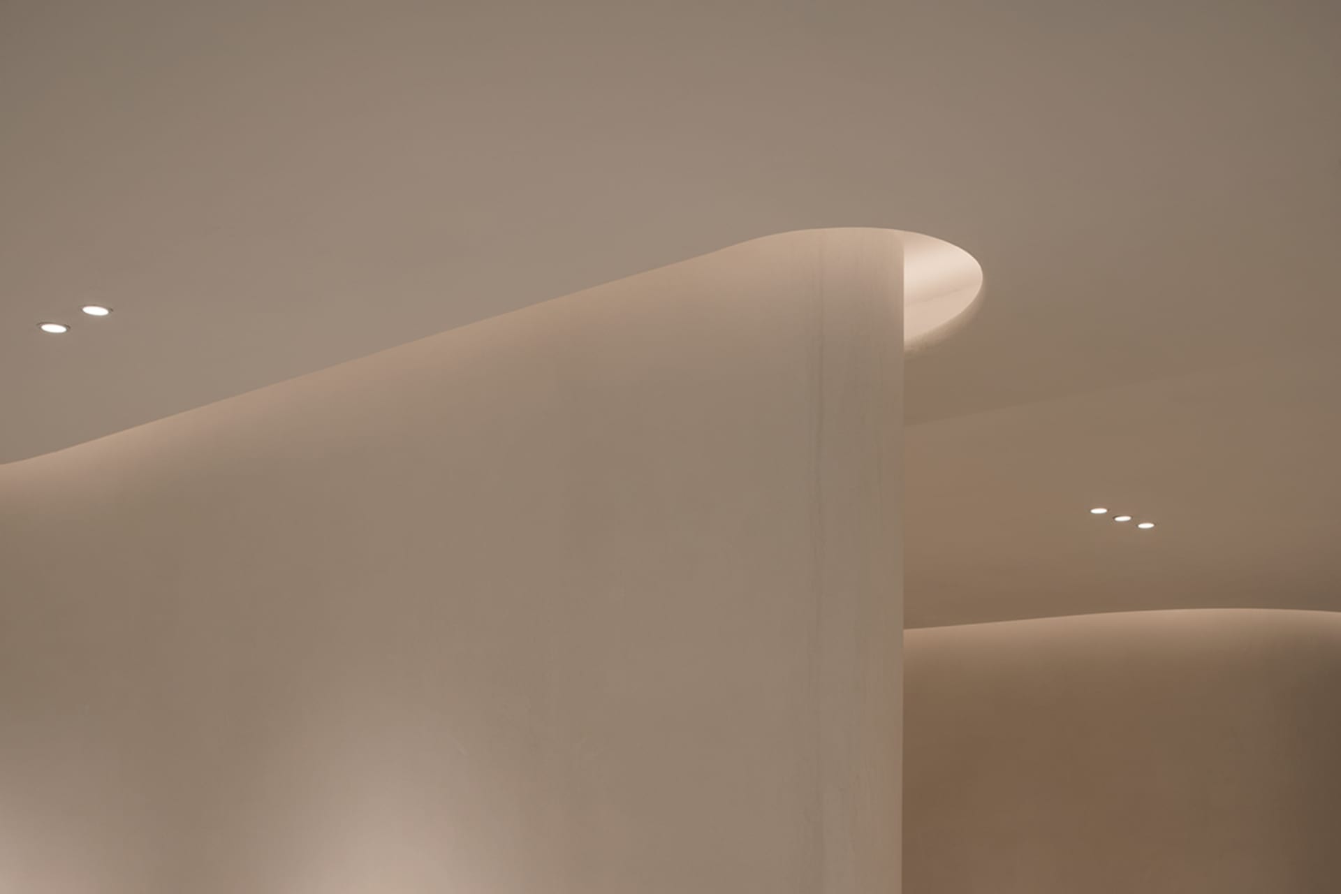 A minimalist architectural ceiling featuring curved white surfaces and modern lighting fixtures.