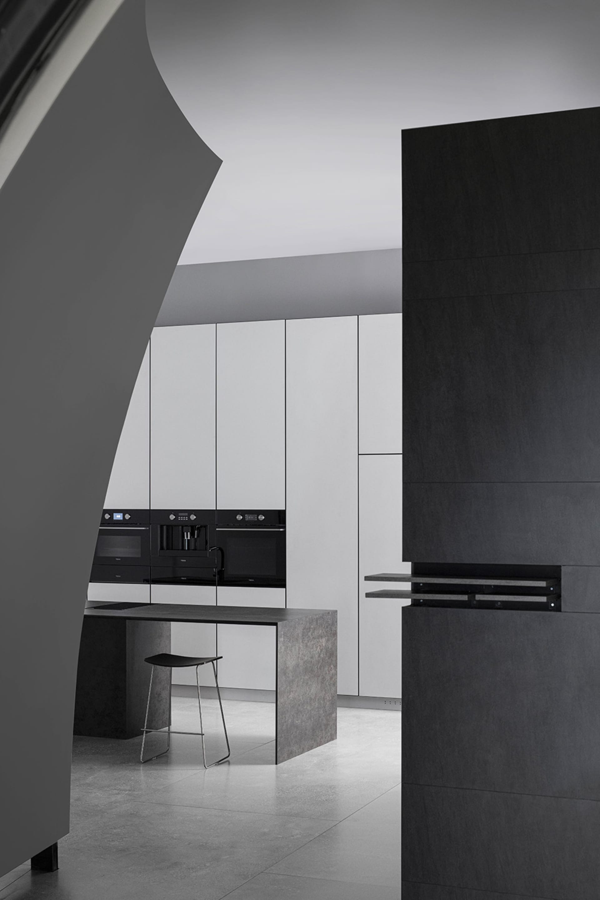 A modern, minimalist kitchen with white cabinets, concrete countertops, black appliances, and a black and metal bar stool.