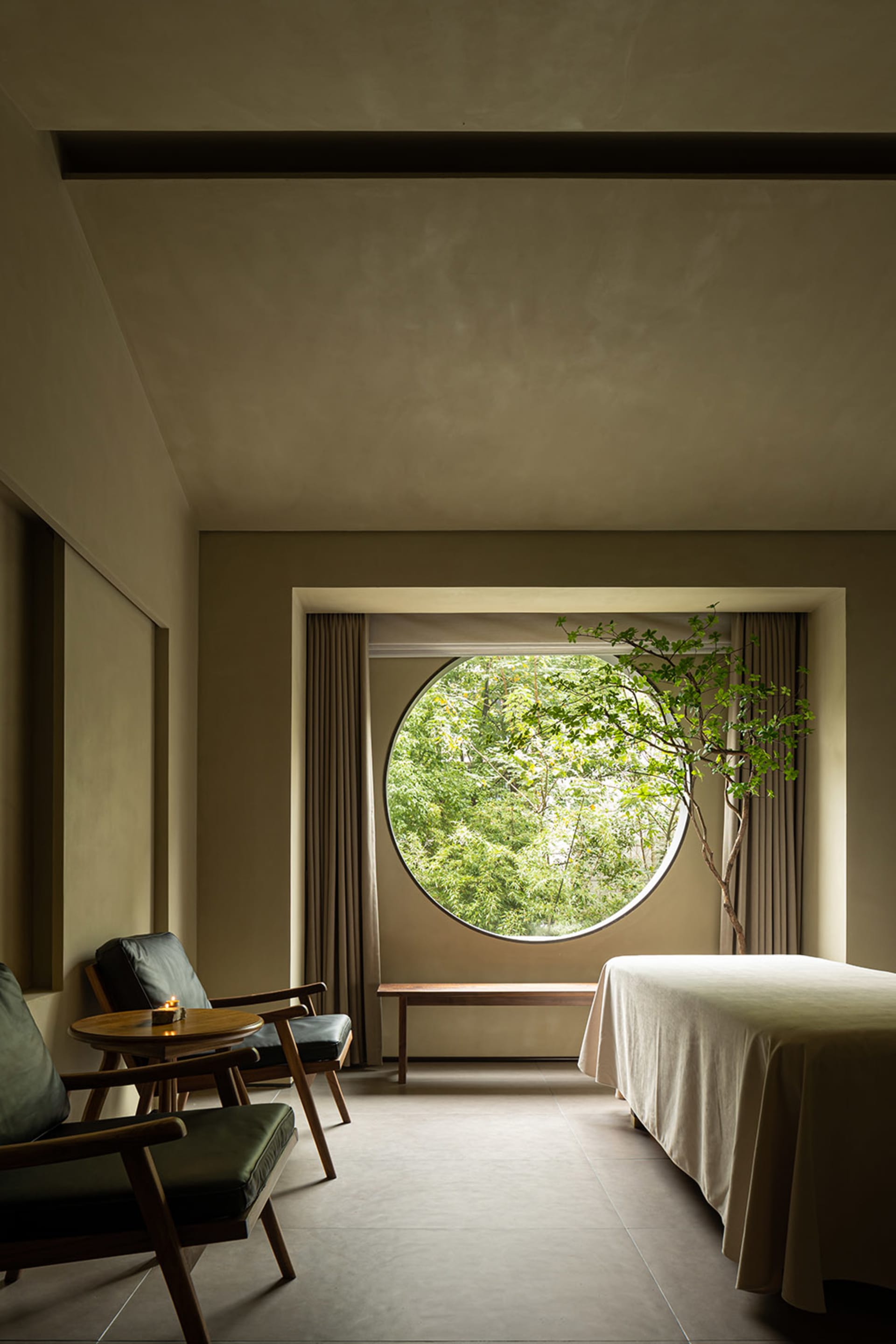 A minimalist bedroom with a round window, textured cloth bedding, and a small tree.