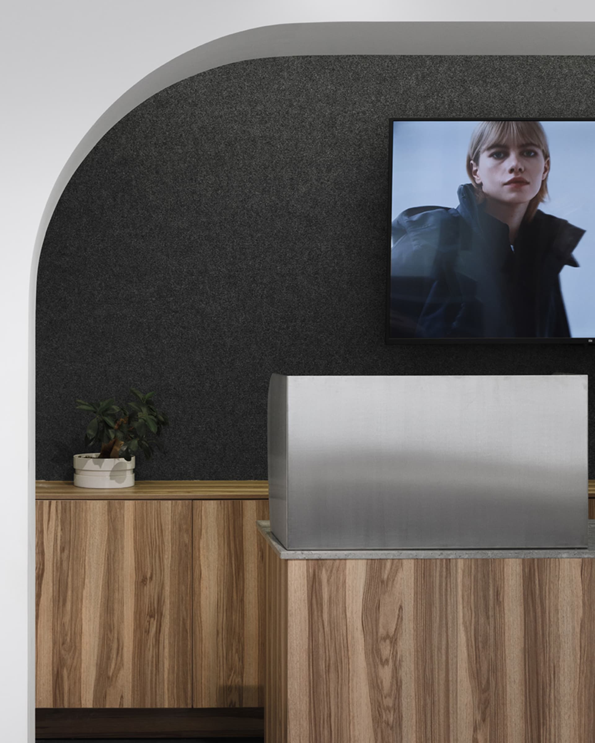 Modern office reception area with a wooden desk, metallic laptop cover, and a TV displaying a woman in a jacket.