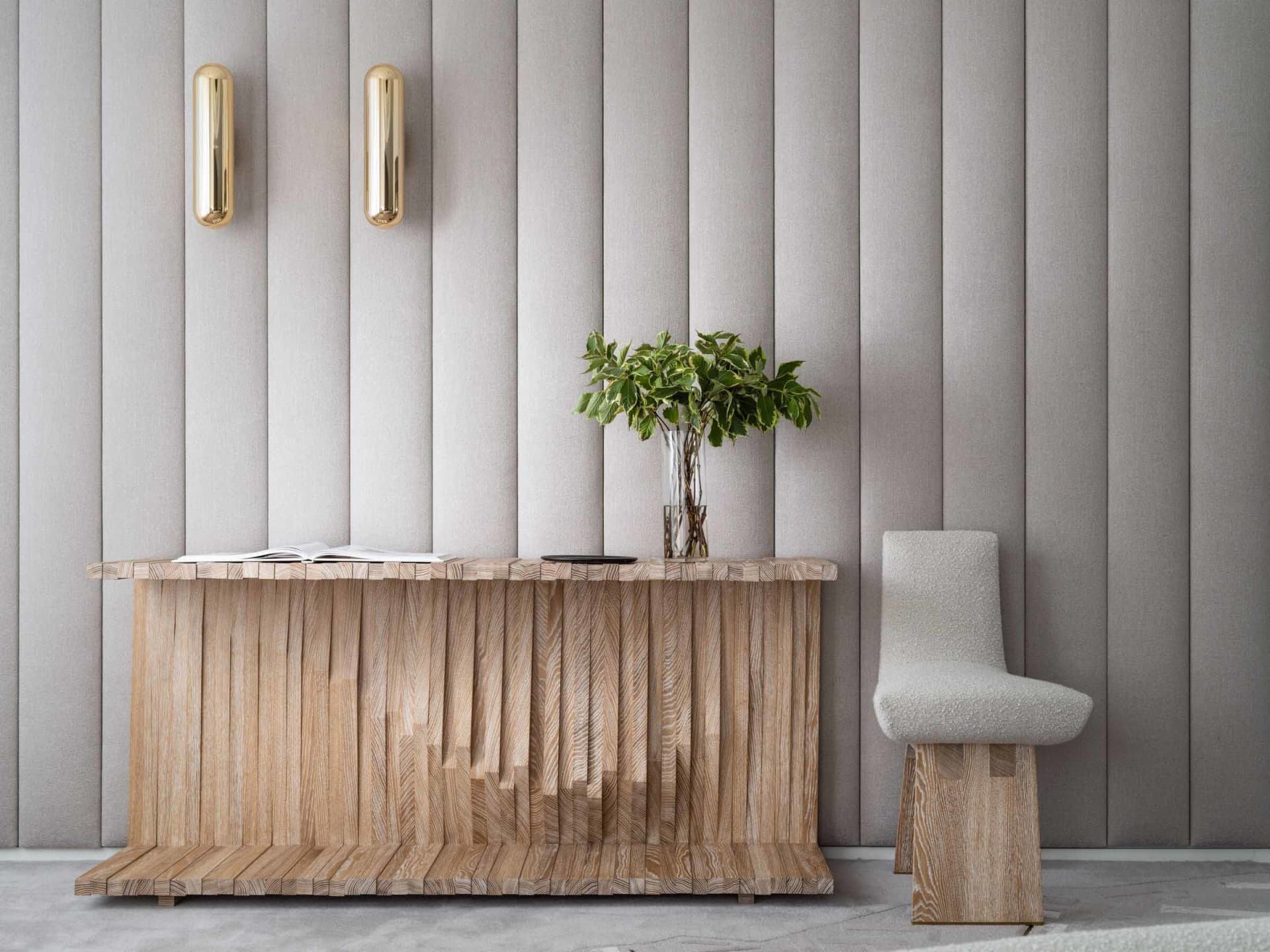 A modern entry console table made of wooden slats with a vase of green plants and a book on top, next to a textured white cha