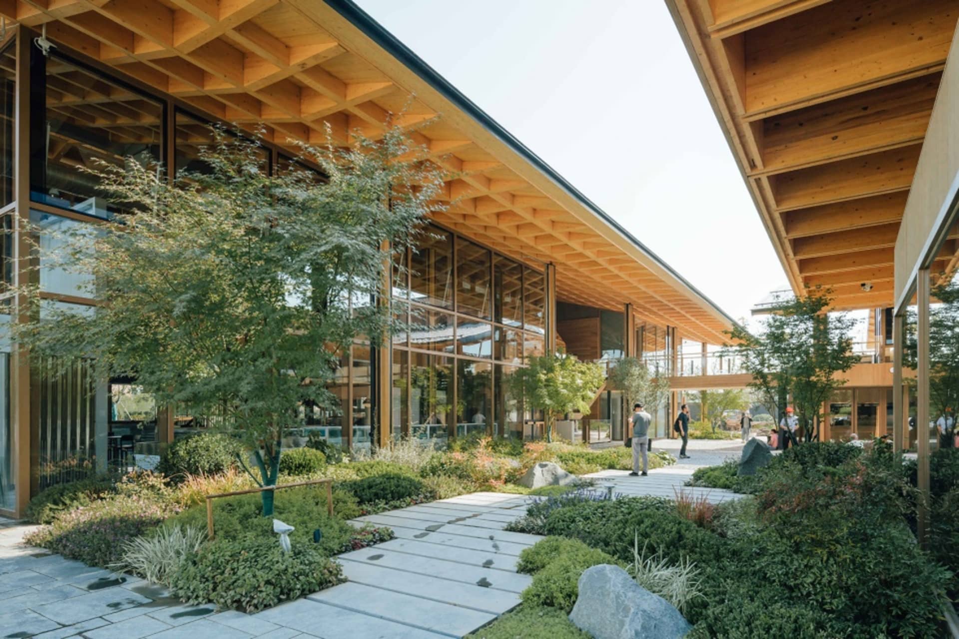 A modern cultural center featuring a wooden facade, large glass windows, and stone elements, surrounded by greenery and pathw