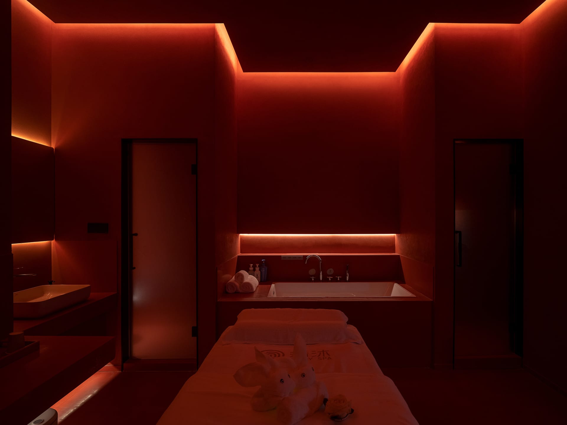A spa room featuring a massage table with plush towels and a bathtub under dim red lighting.