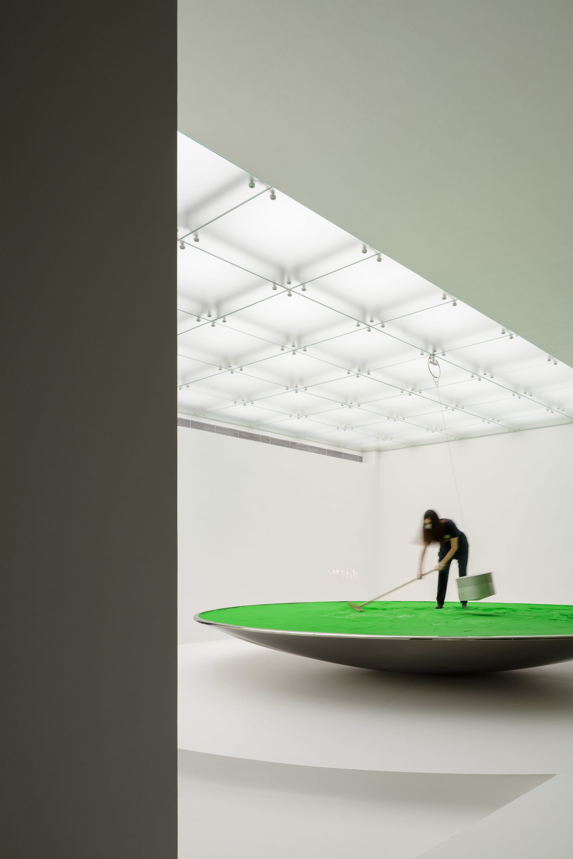A minimalist art installation featuring a green surface being raked within a metallic bowl.