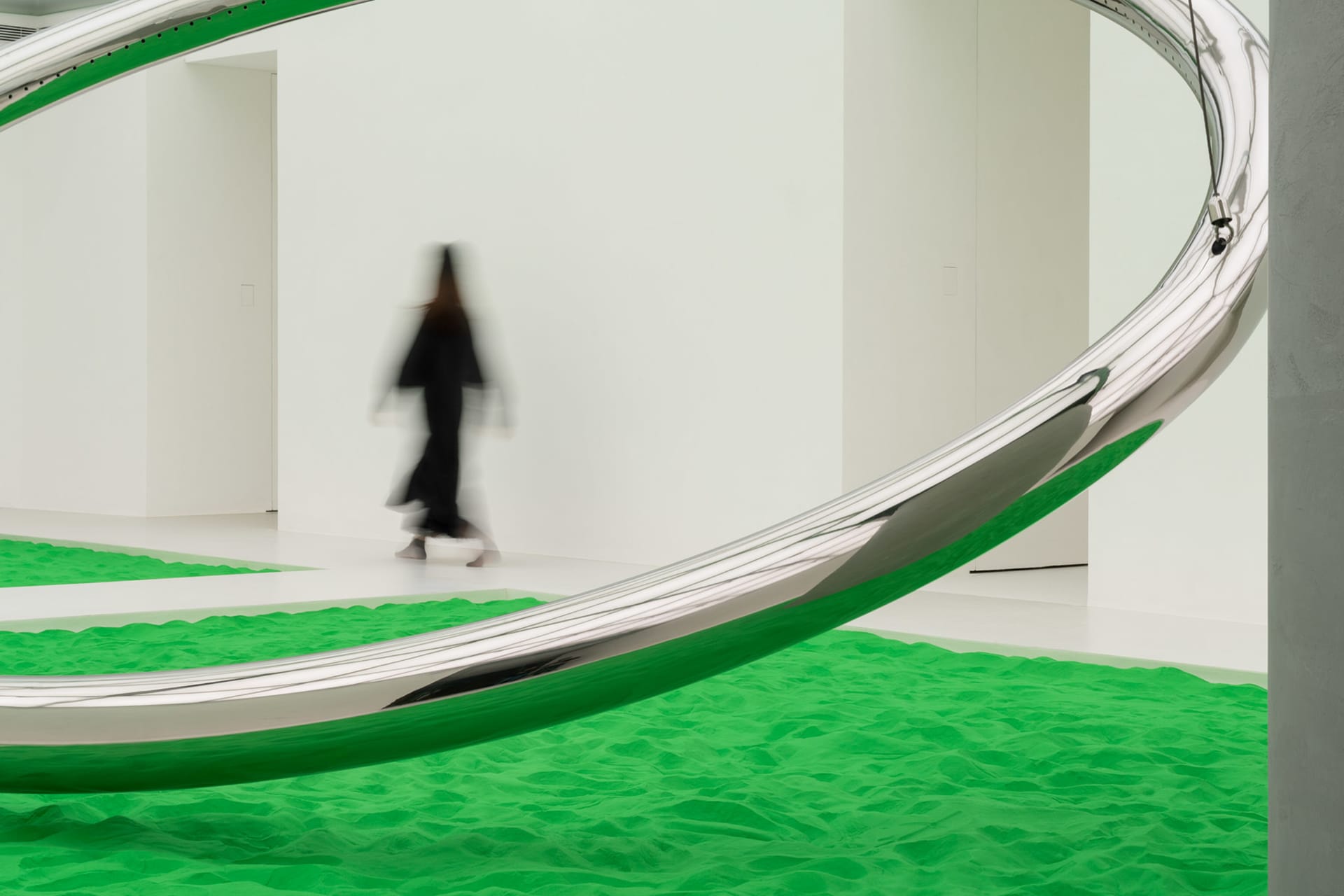 A modern art installation featuring a large, curved metallic structure and green sand floor.