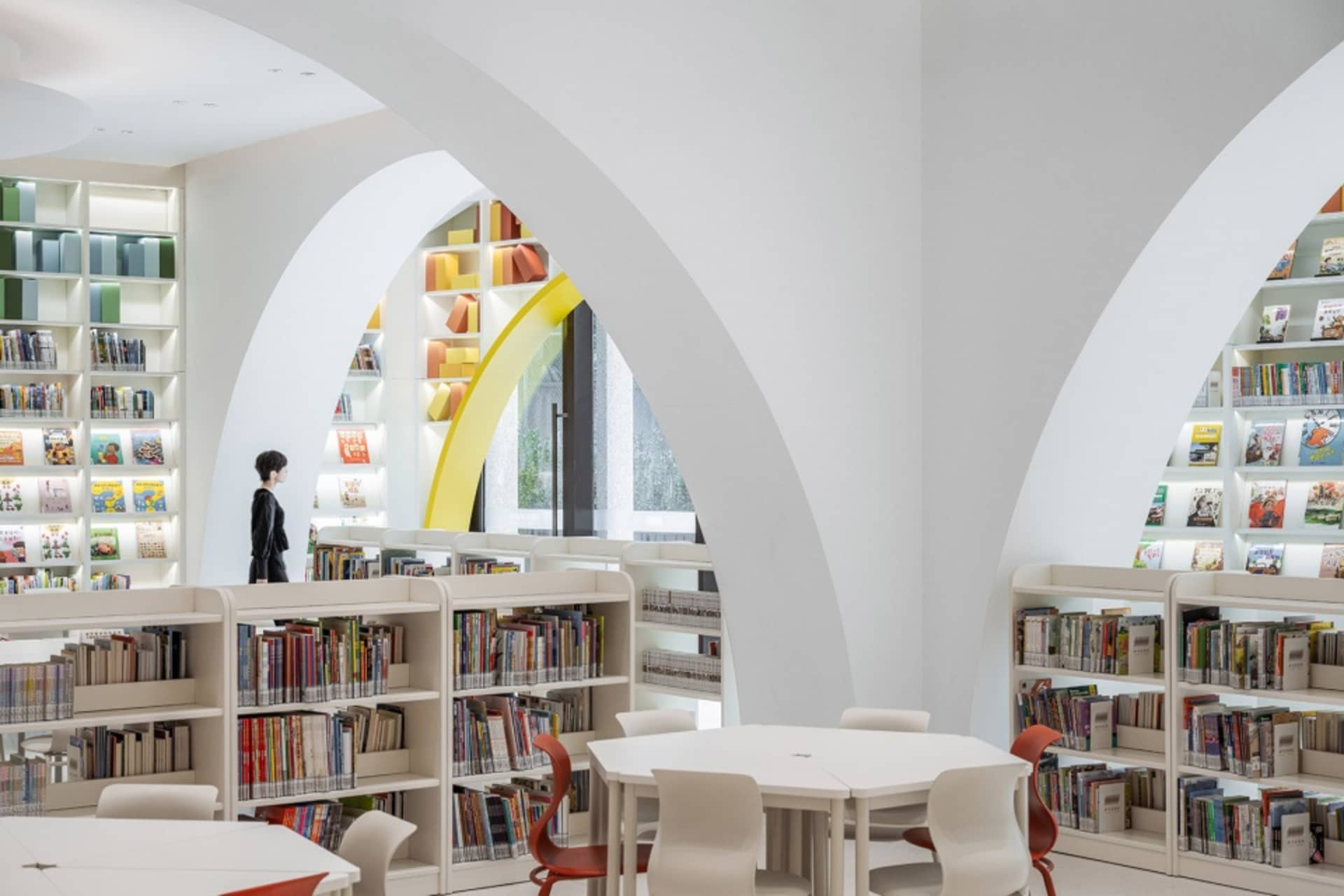 A modern library interior with white-painted walls, circular arches, and colorful bookshelves.