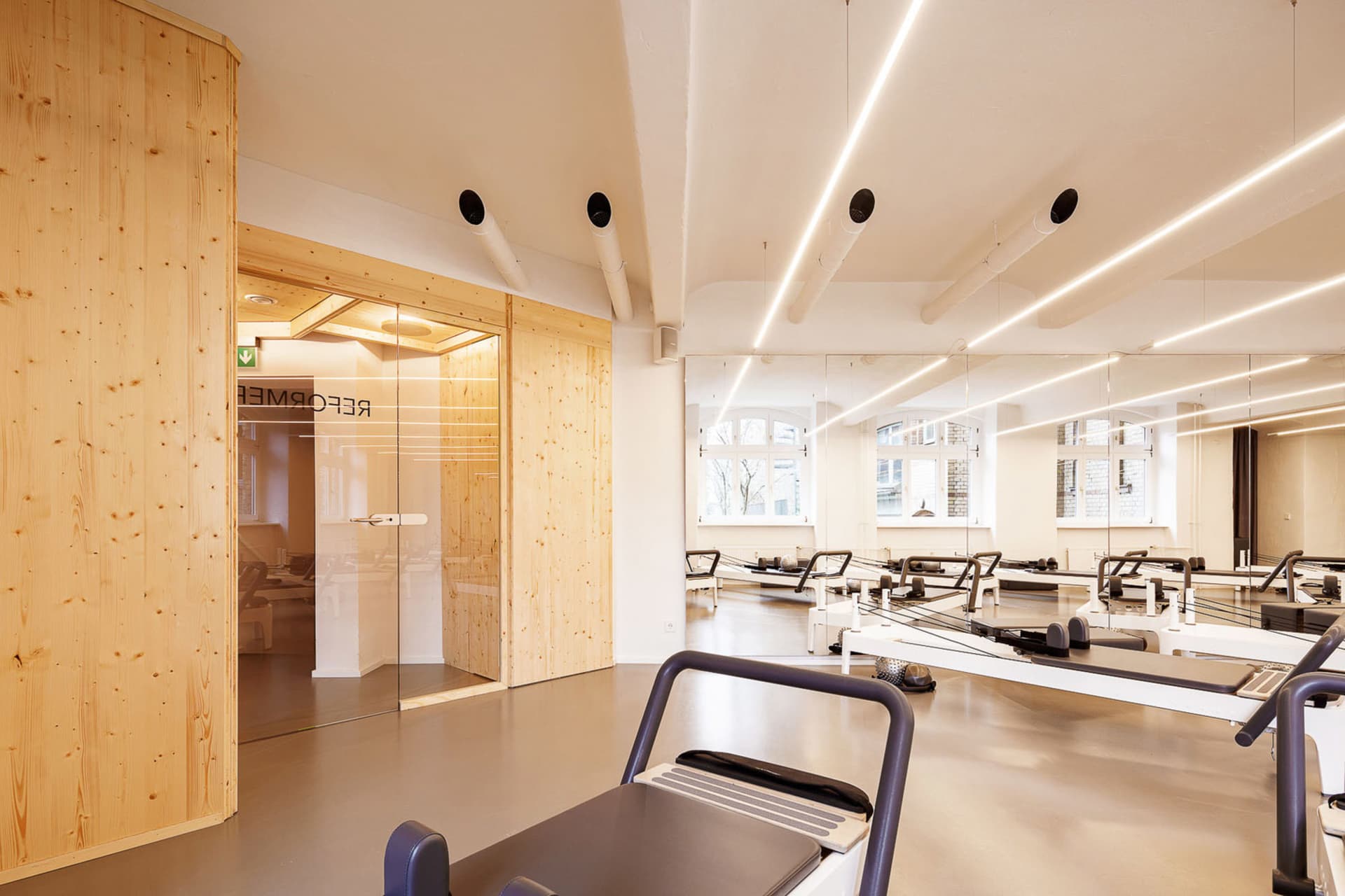 A minimalist fitness studio with wooden walls and floors, featuring Pilates reformers and mirrors.