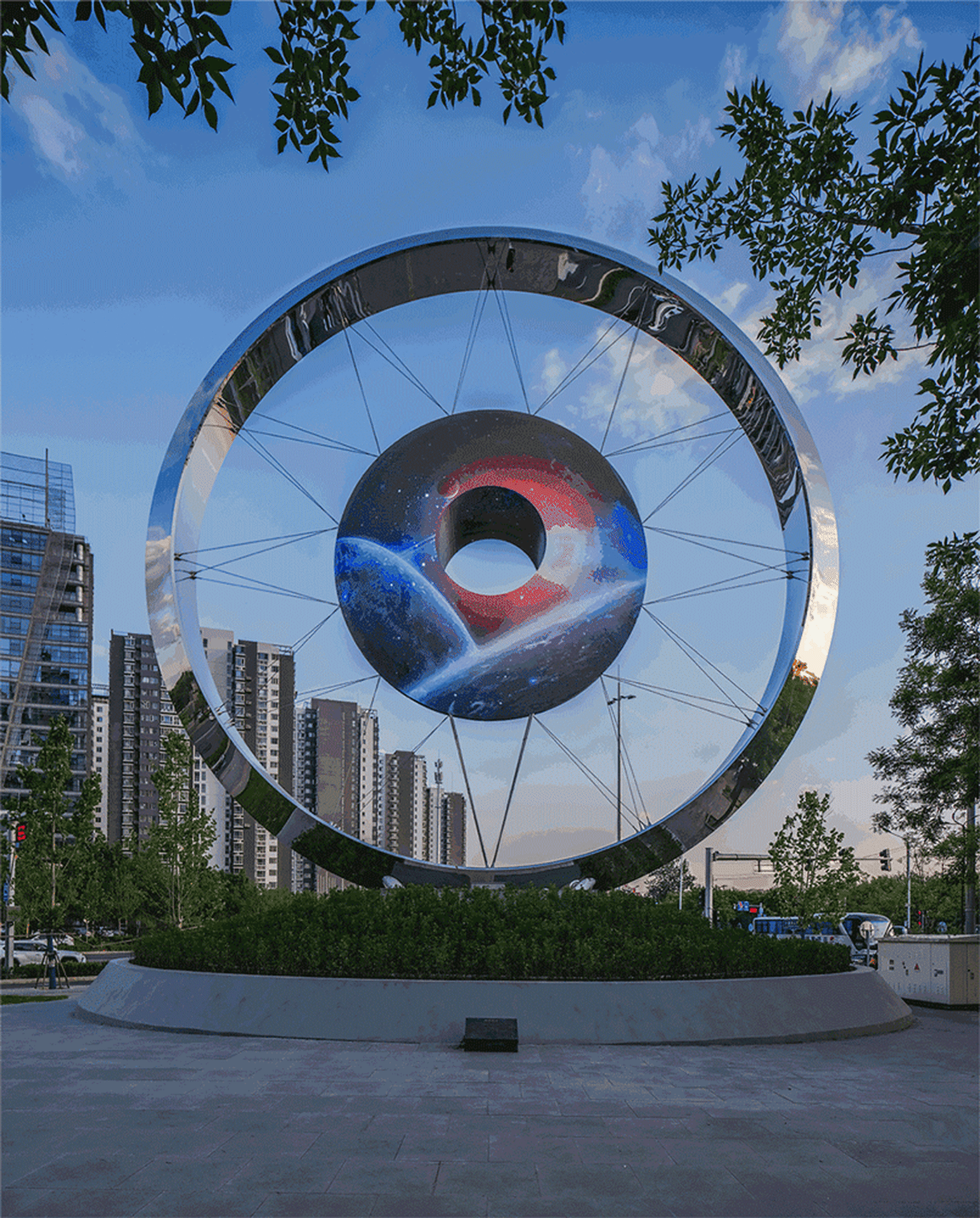 A large metallic sculpture with a galaxy-themed central ring located in an urban city square.