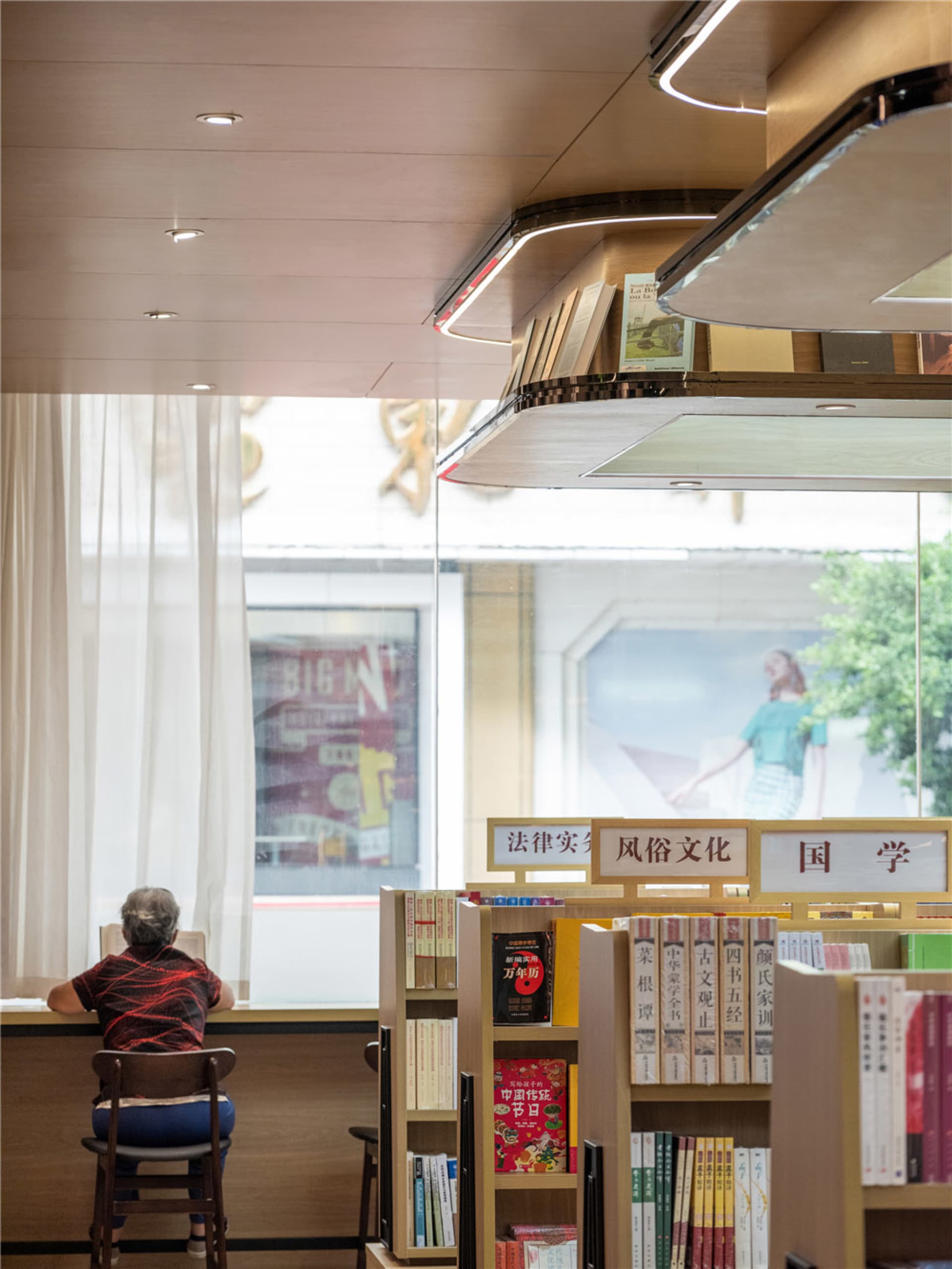 A modern library with a wooden staircase, white curtains, and bookshelves filled with books.