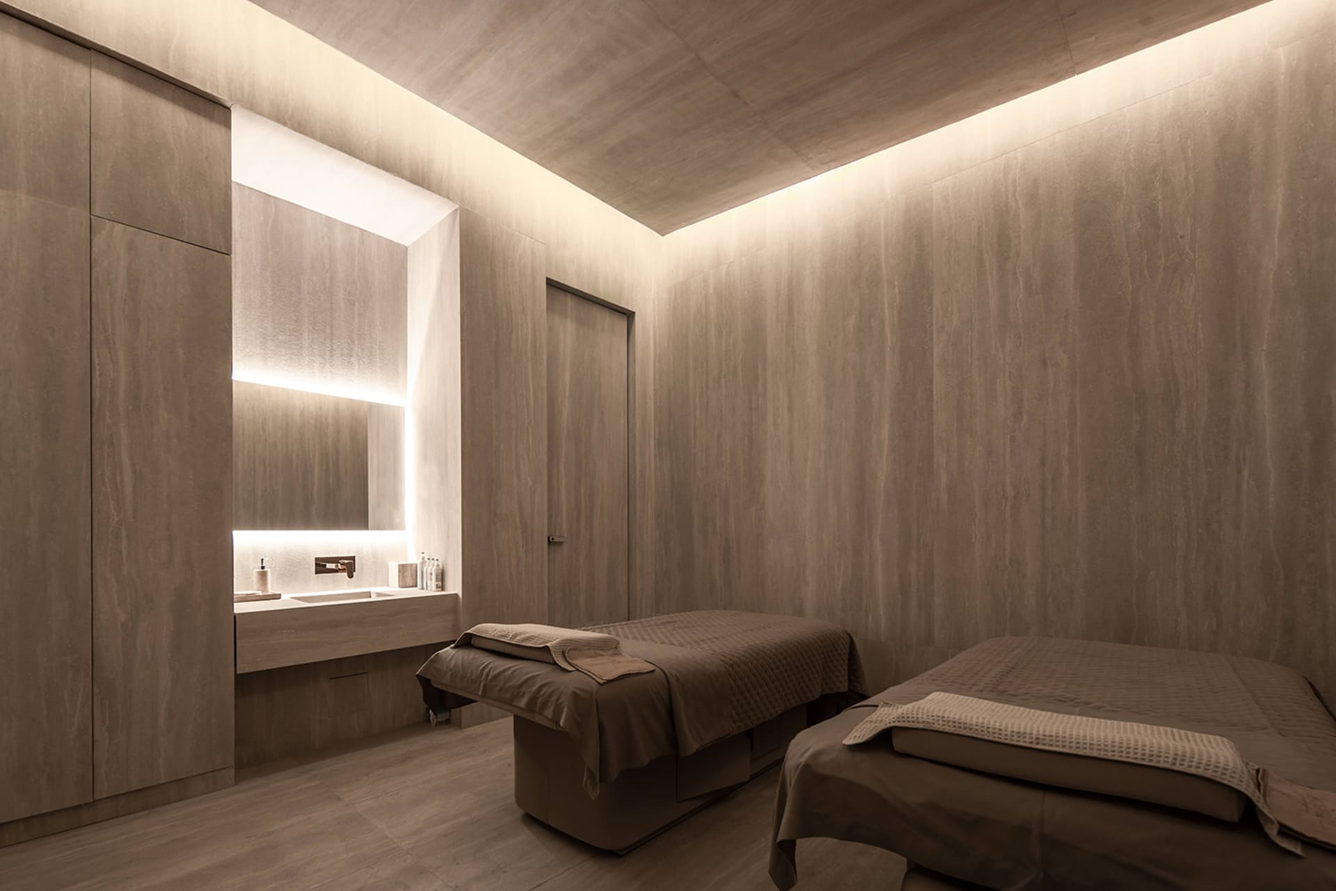 A spa treatment room with concrete walls, two massage tables, and a sink area with ambient lighting.