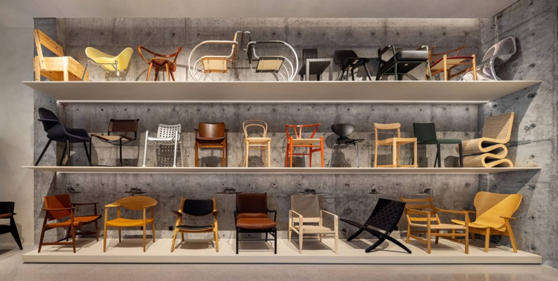 A collection of industrial design chairs displayed on two shelves with a concrete background.