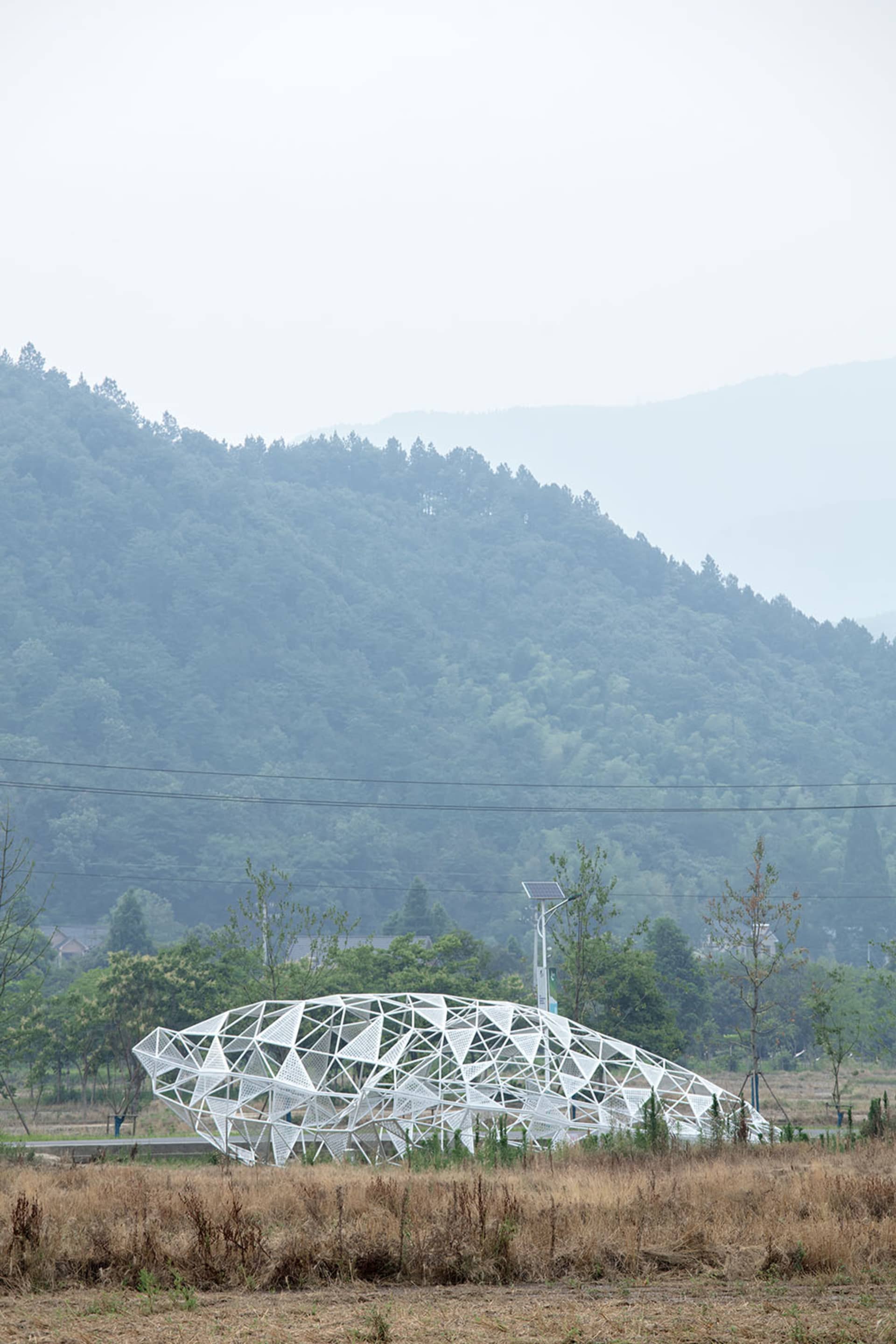 A metallic, modern structure resembling a wave or organic form, located in a field with a mountainous backdrop.