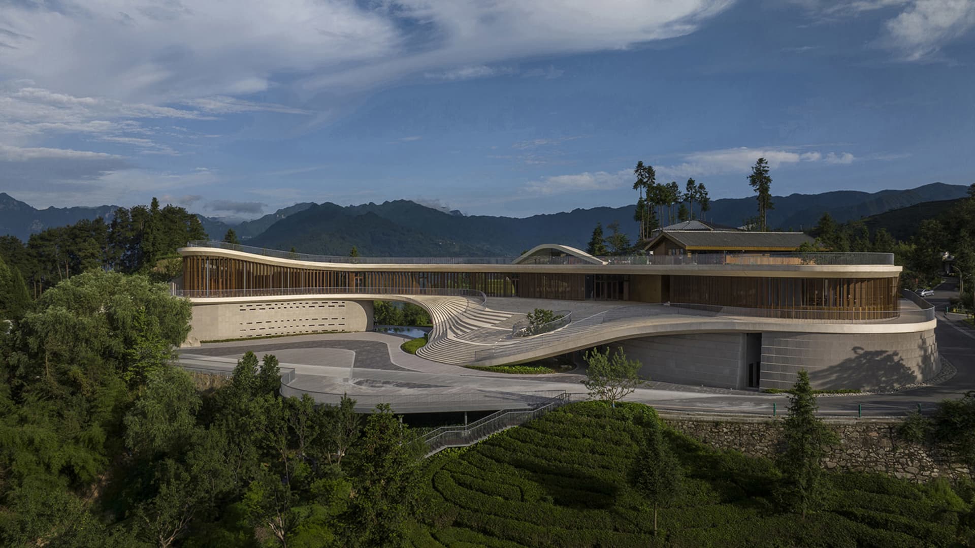 A modern hotel with concrete and glass exterior, surrounded by greenery and mountains.