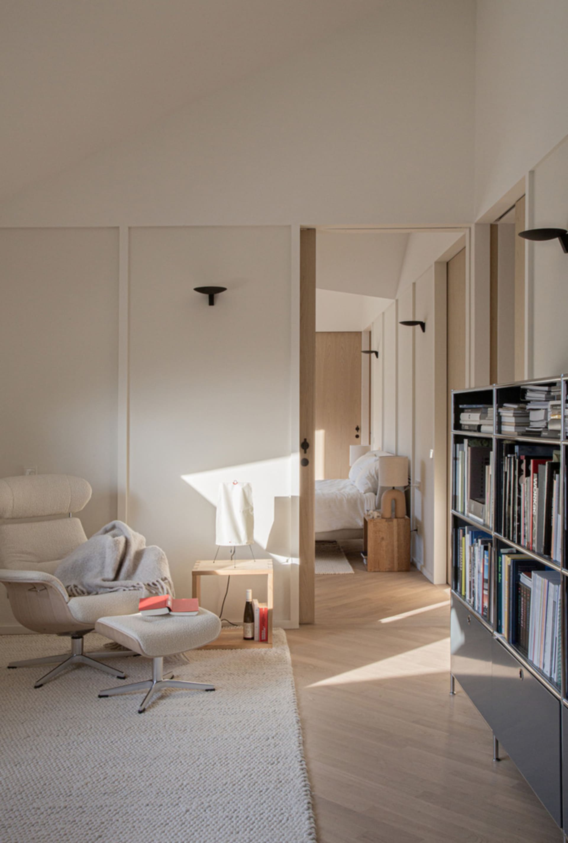 A minimalist living room with white wood furniture and a white rug, featuring a comfortable seating area and a bookshelf.