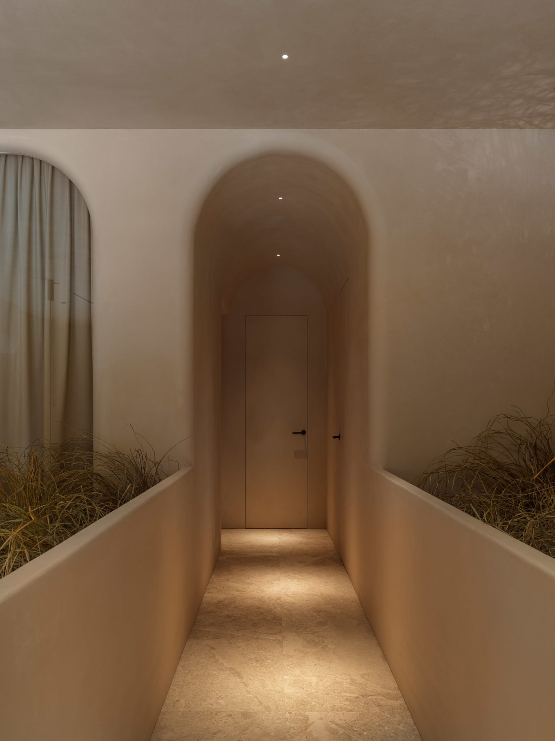 A minimalist corridor featuring a concrete floor and walls, with a single door at the end and small plants on either side of