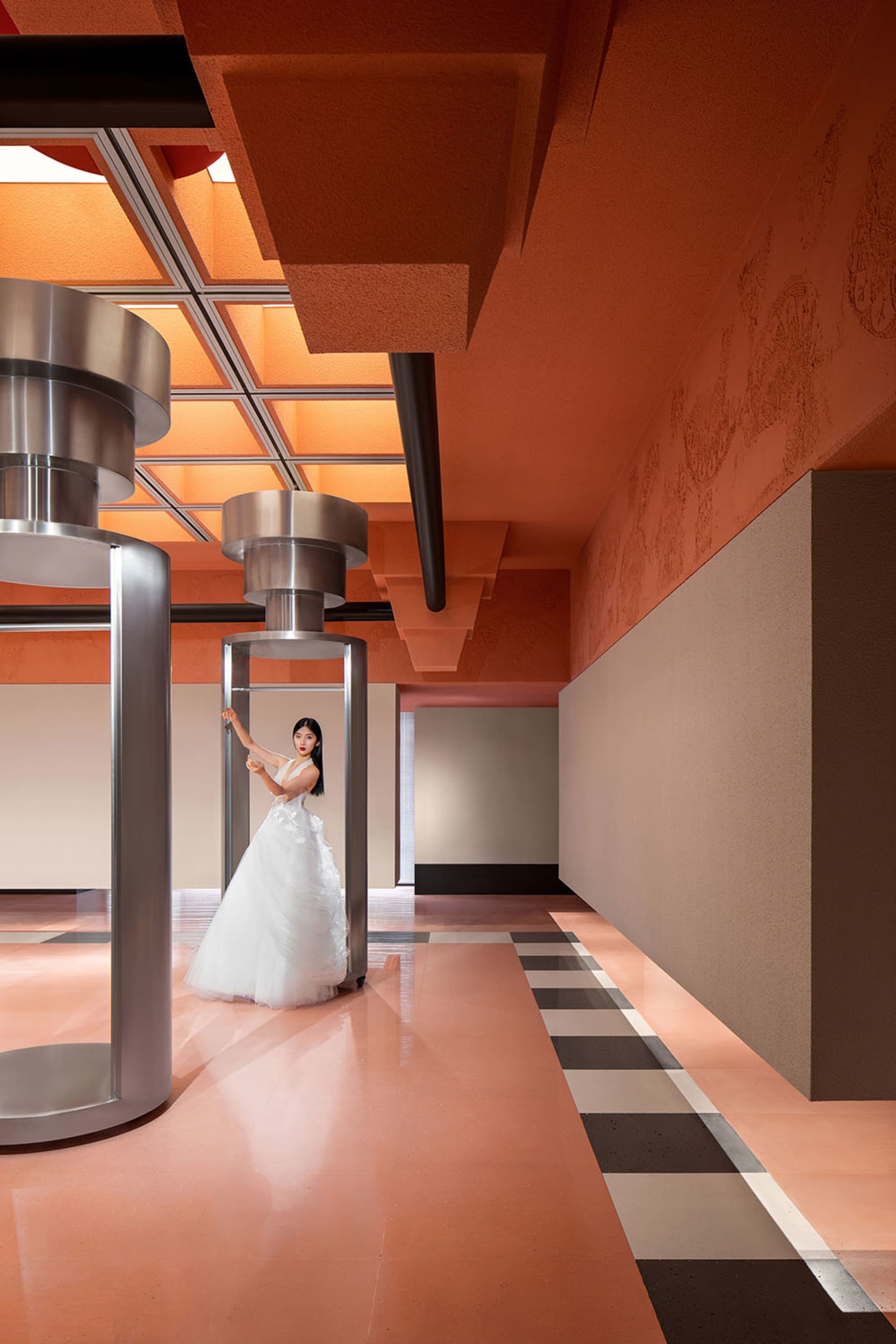 Fashion shoot featuring a woman in a white dress standing in a modern, industrial space with metallic pillars and a geometric