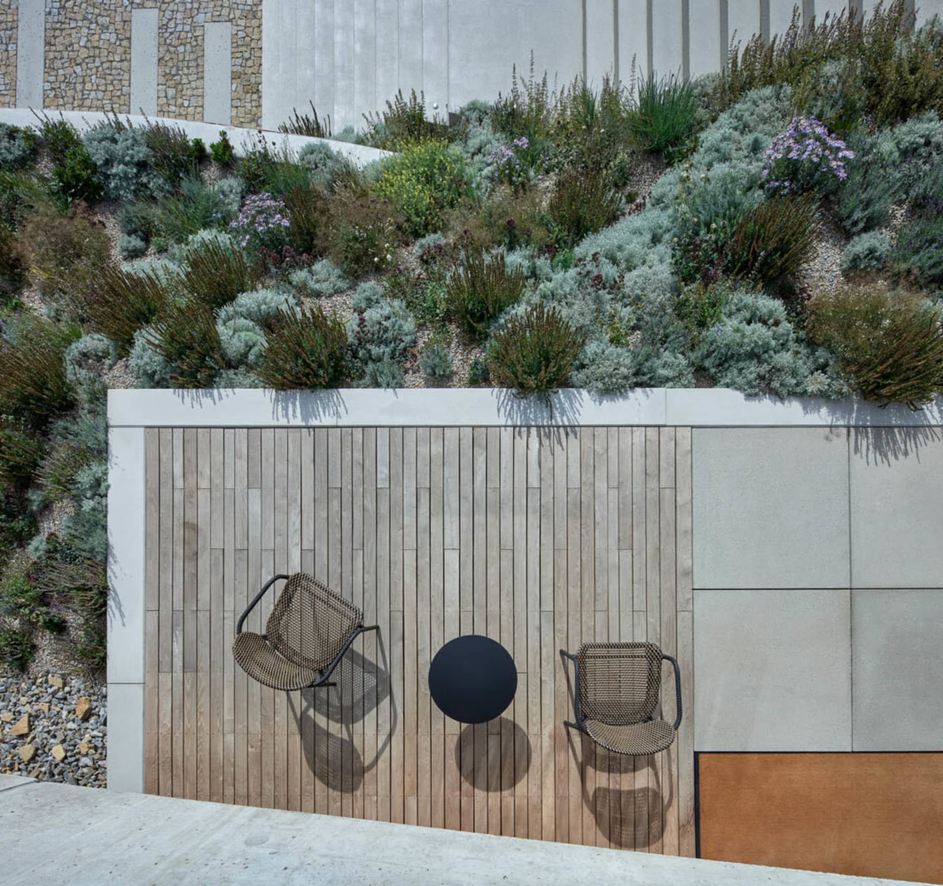Top view of a modern garden with a wooden deck, concrete walls, and rocks, featuring a circular table and two metal chairs.