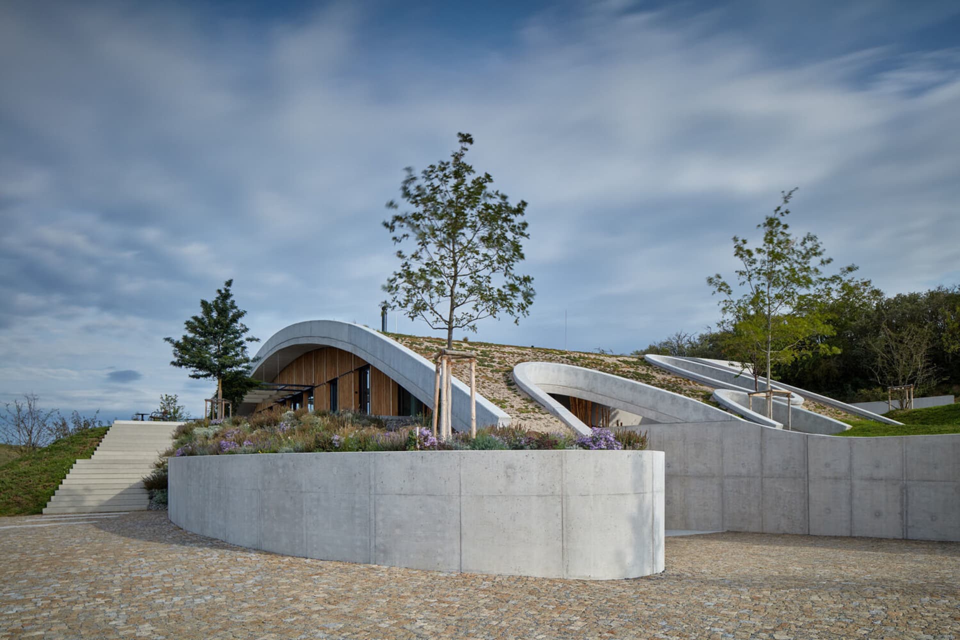 A modern building constructed with concrete and wooden elements, featuring curved architectural elements and a landscaped for