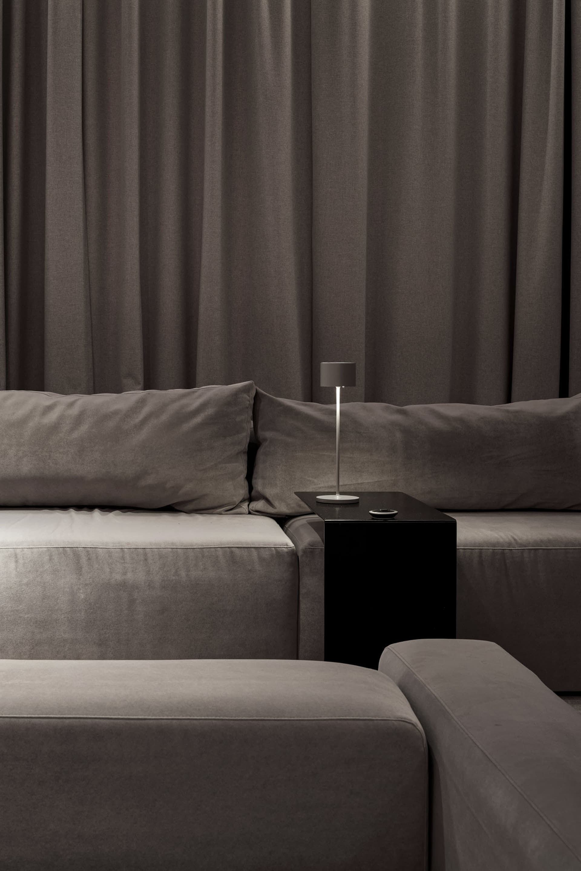 Minimalist living room with a large, comfortable-looking grey velvet couch, complemented by neutral-toned pillows and curtain