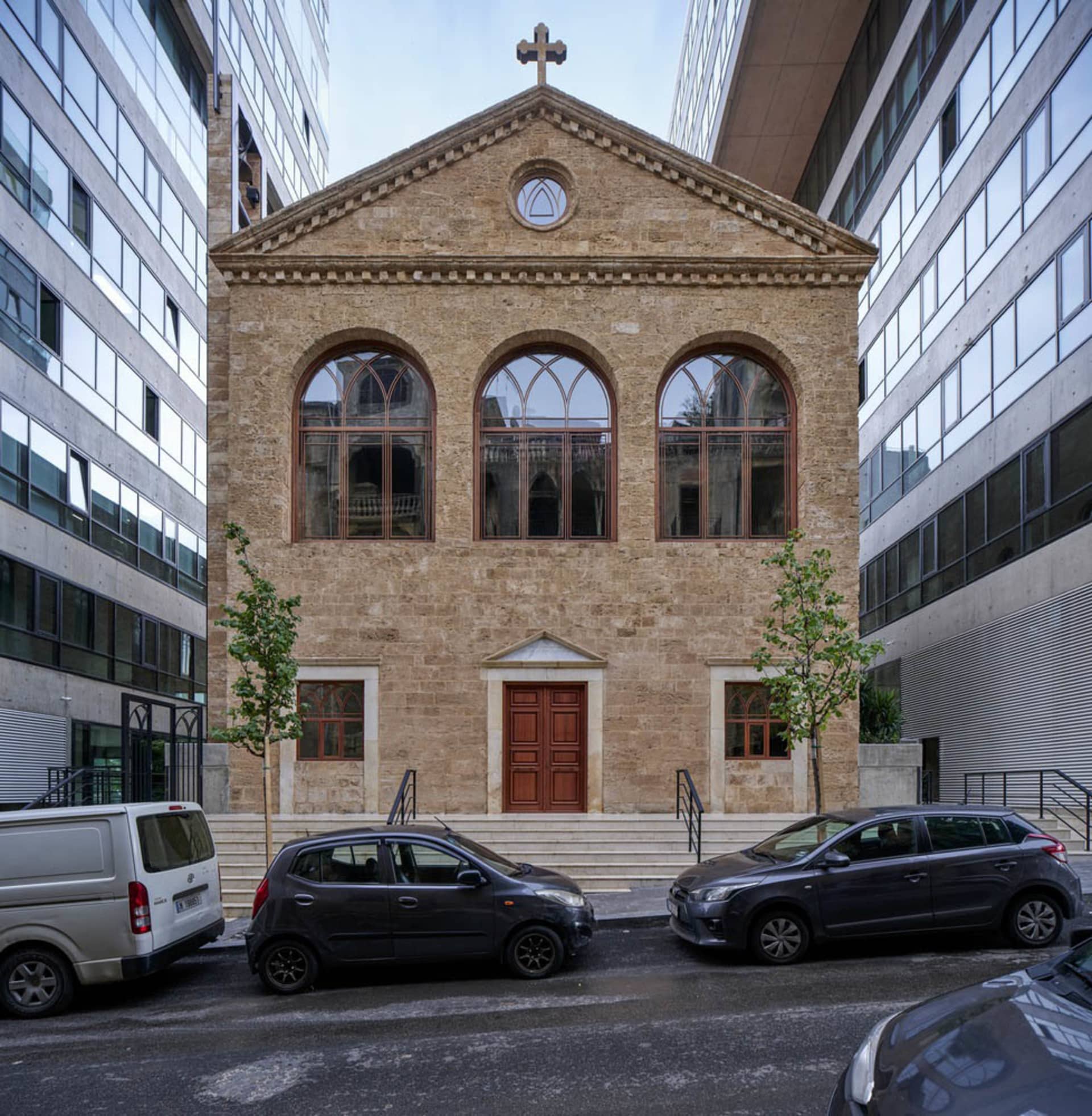 A modern stone church with three arched windows and a cross on the roof, surrounded by modern buildings and parked cars.