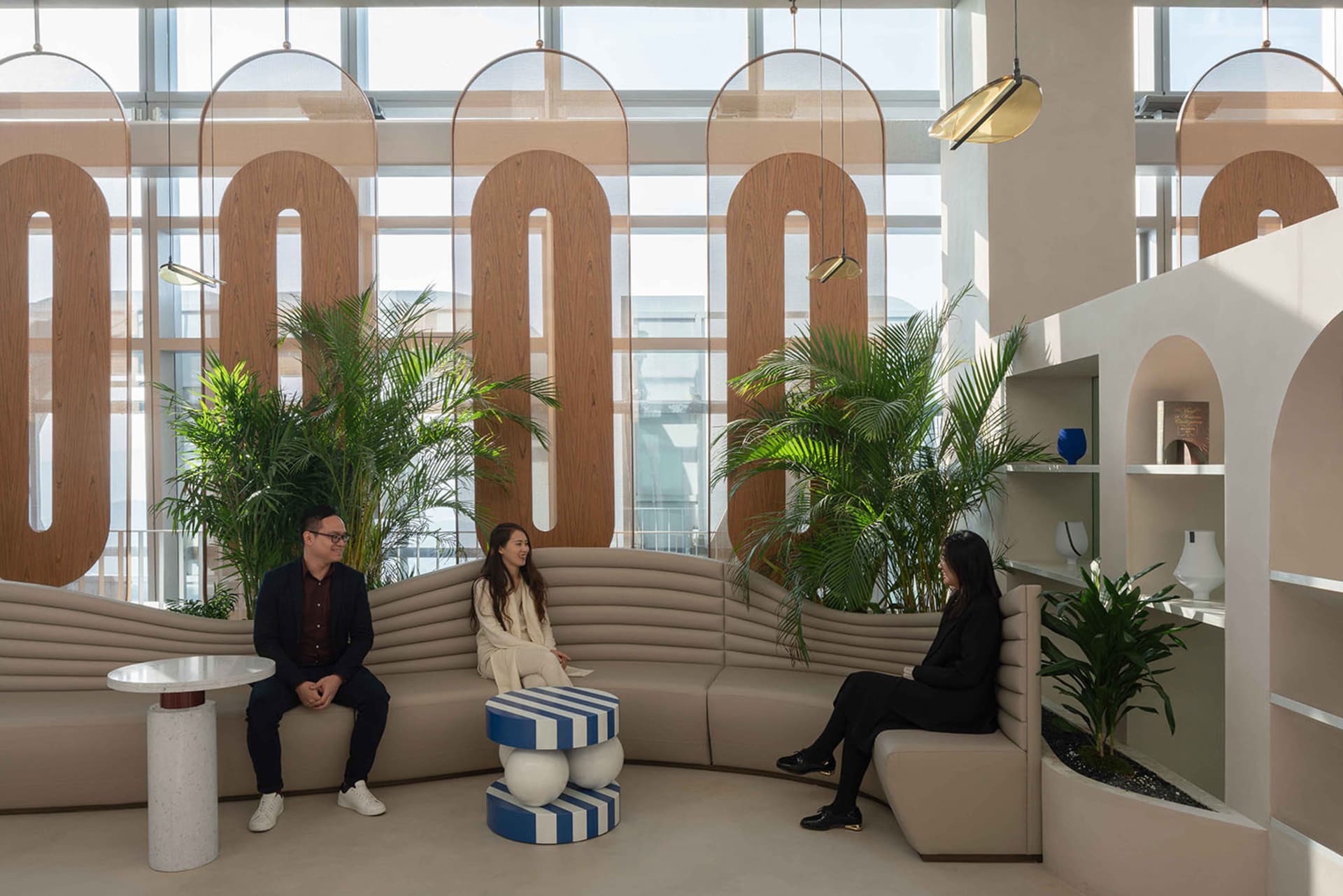 People sitting in a modern office lounge with concrete and fabric seating, glass partitions, and green plants.