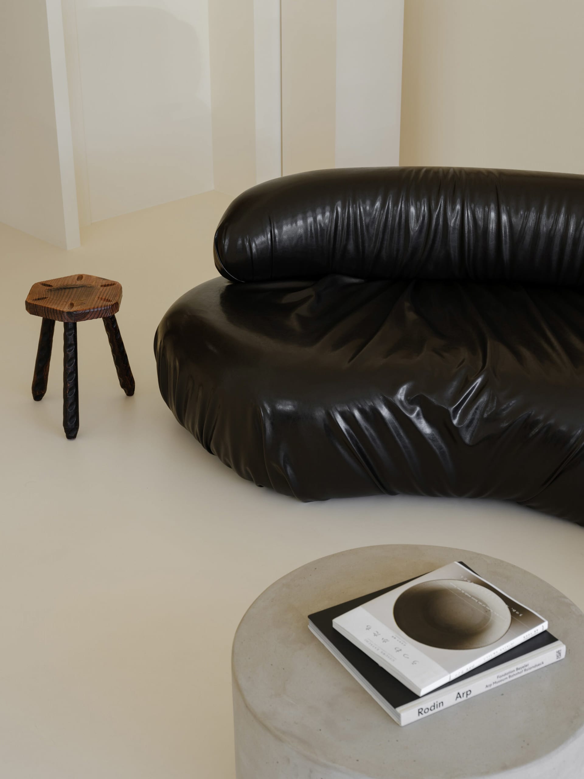 A minimalist black leather sofa in a living room setting, next to a vintage wooden stool and a concrete table with books.
