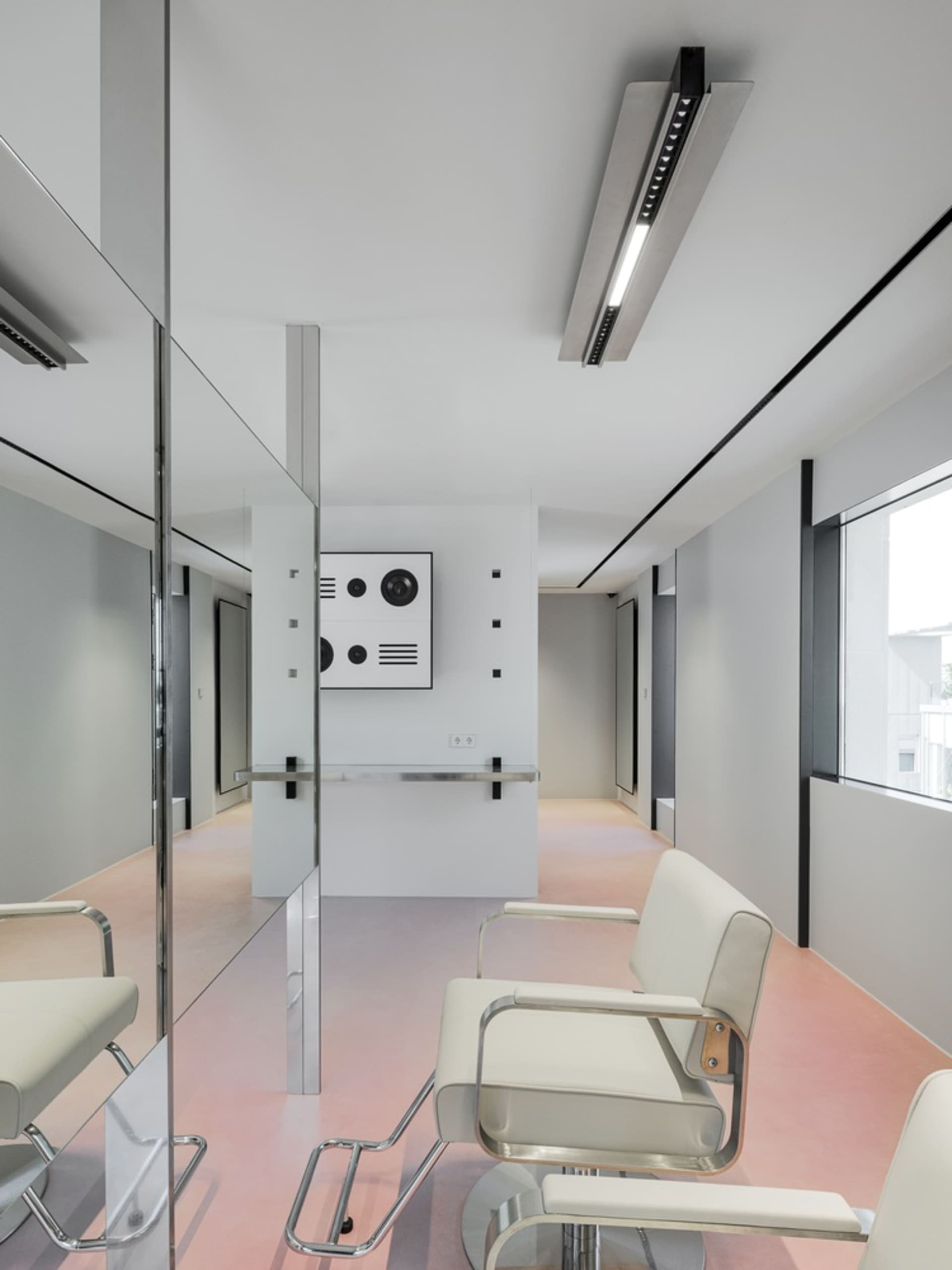 A minimalist white interior of a hair studio, featuring sleek modern chairs, a counter with a mirror, and large windows allow