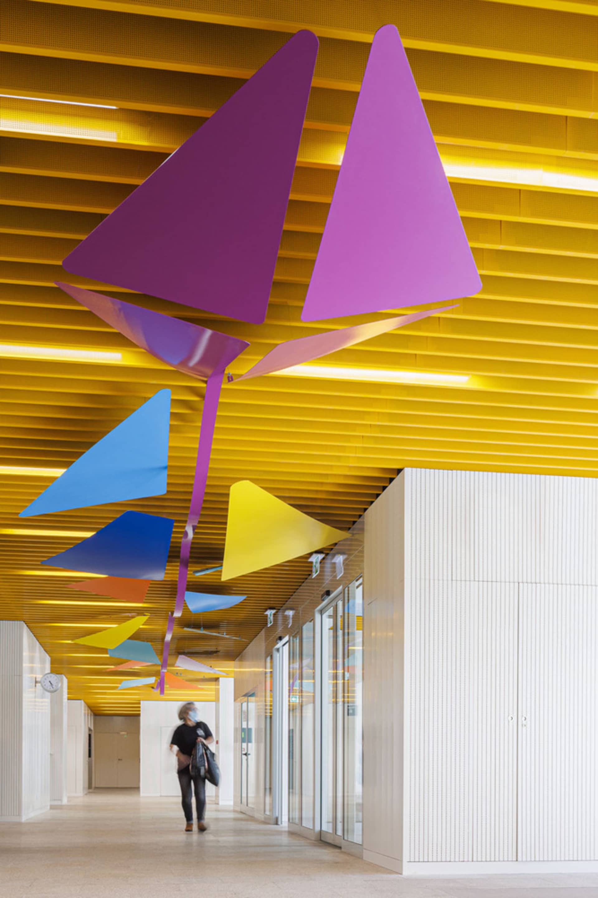 A stylized modern interior corridor with colorful geometric shapes hanging from the ceiling, featuring a geometric metal art
