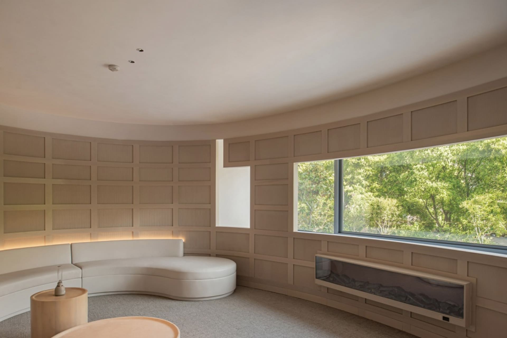A modern interior space with neutral tones, featuring a curved sofa, a large window overlooking trees, and a fireplace.