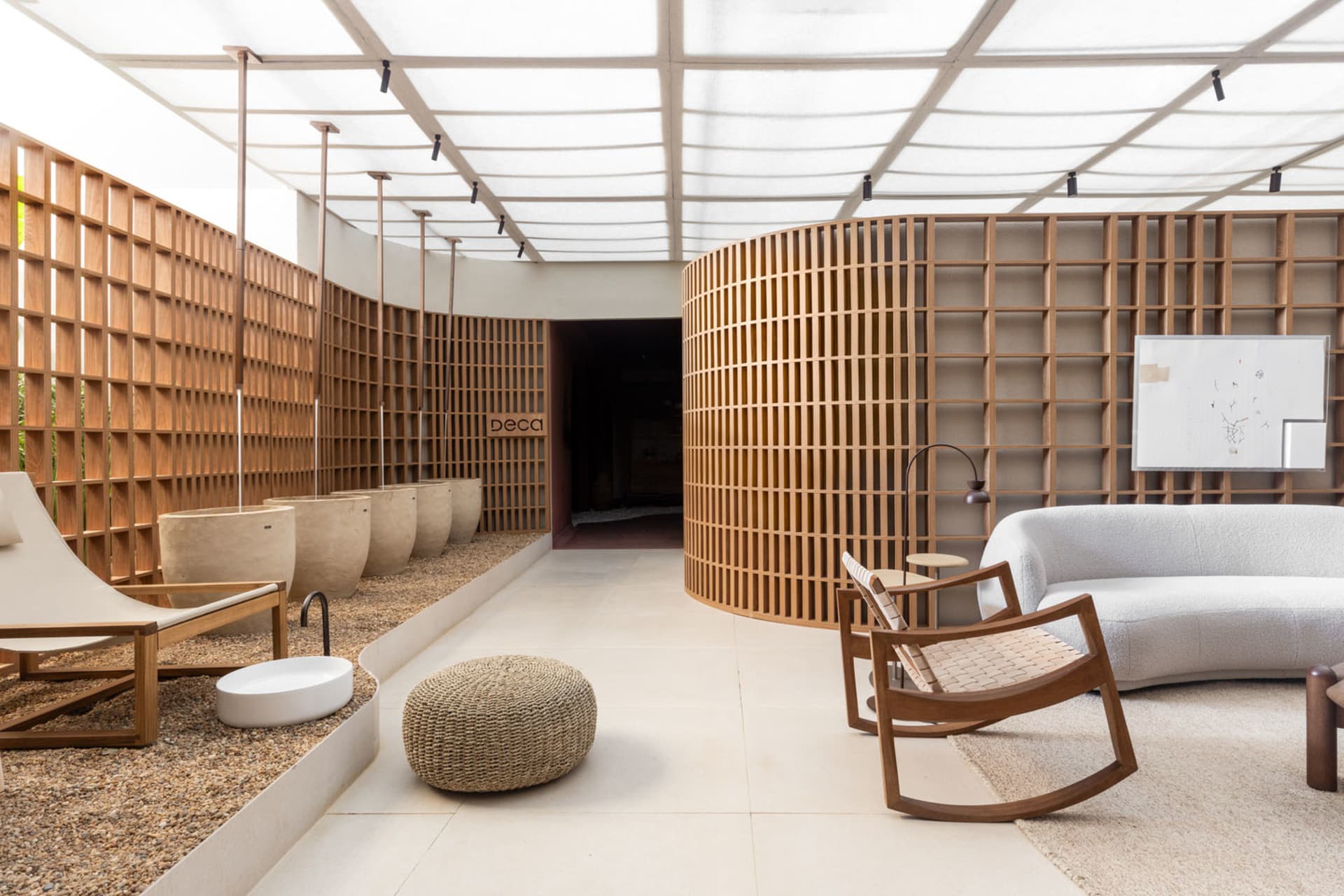 A modern living room with wooden walls and furniture, featuring a white lounge chair, a round pouf, and a curved wooden sofa.