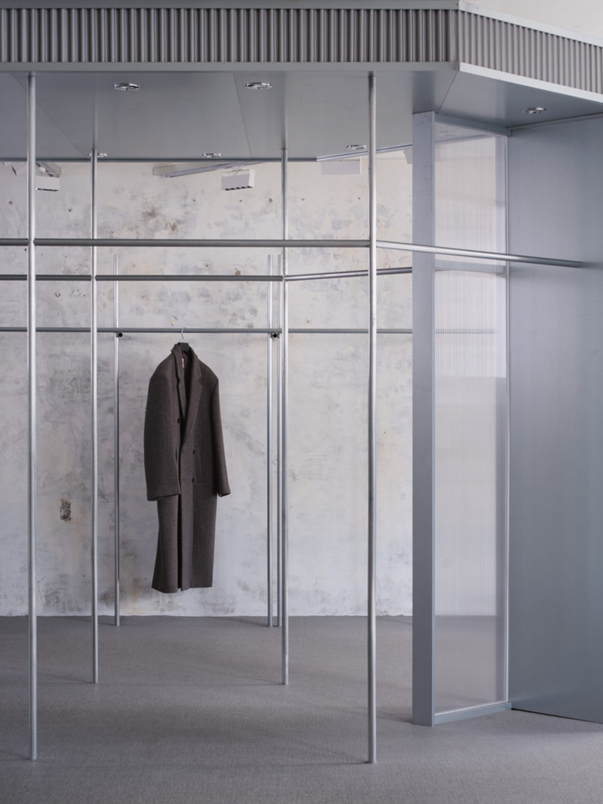 極簡展示間內，一件棕色外套懸掛於金屬架上，搭配玻璃牆與紋理混凝土牆面。