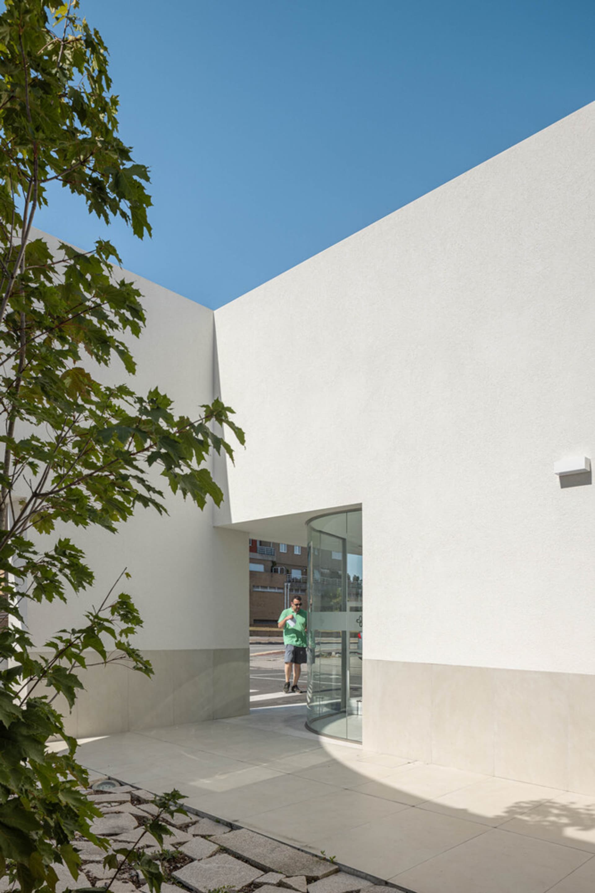 A modern white concrete exterior building with greenery and a clear blue sky in the background.
