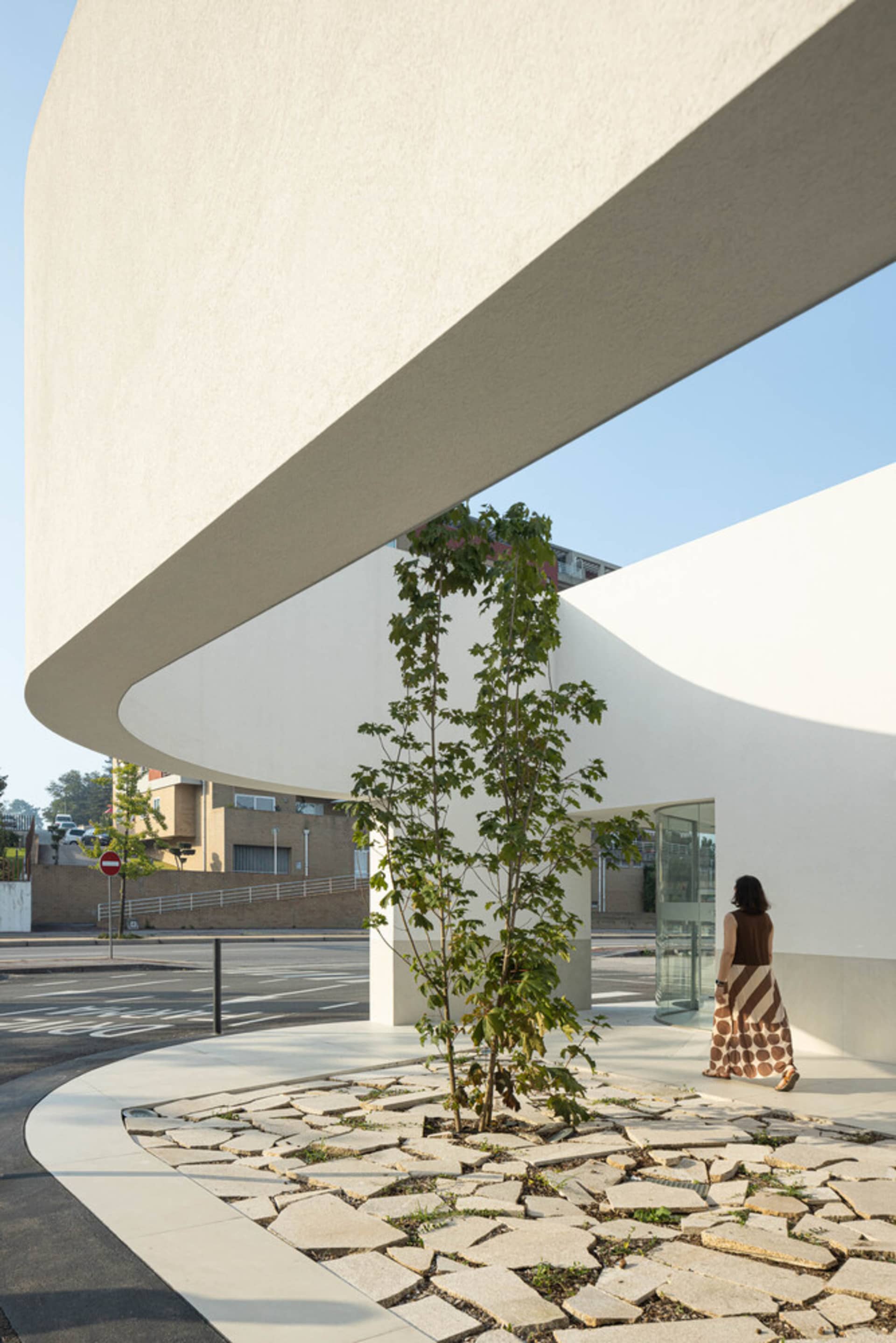 A woman walks past a modern entryway featuring a tree planted in a bed of concrete and glass tiles.