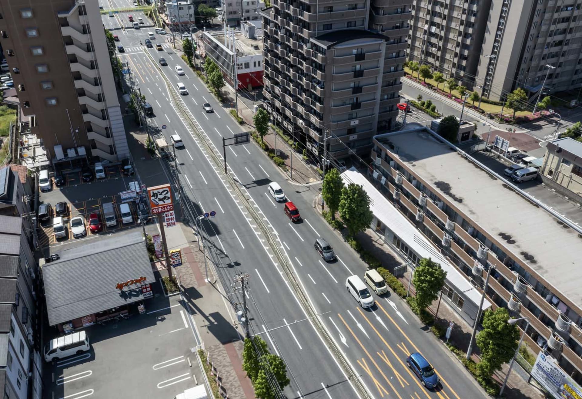 A wide urban road with multiple lanes, surrounded by high-rise buildings and greenery, viewed from an aerial perspective.