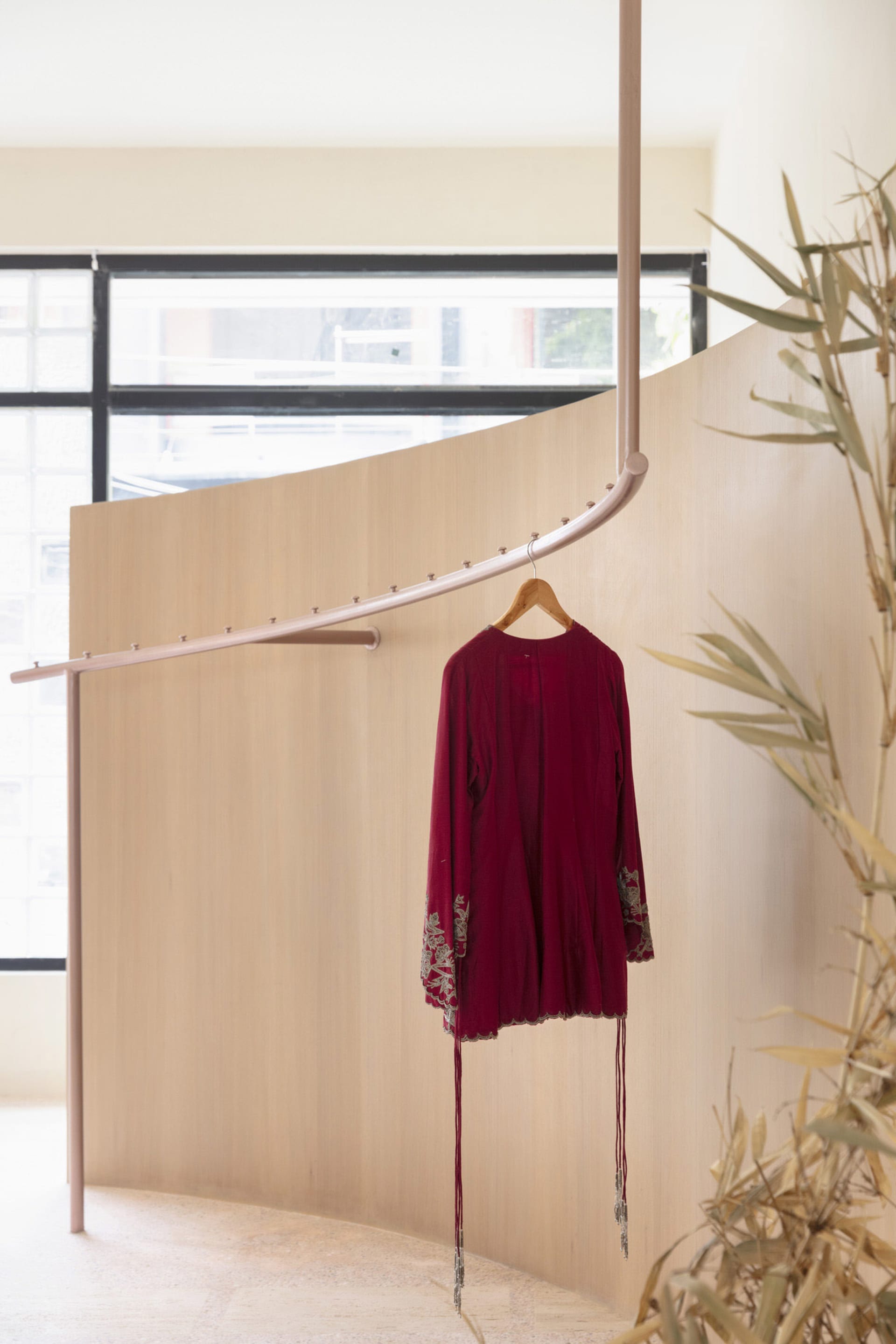 A metallic hanger displaying a burgundy embroidered jacket against a wooden display wall and a leafy plant.
