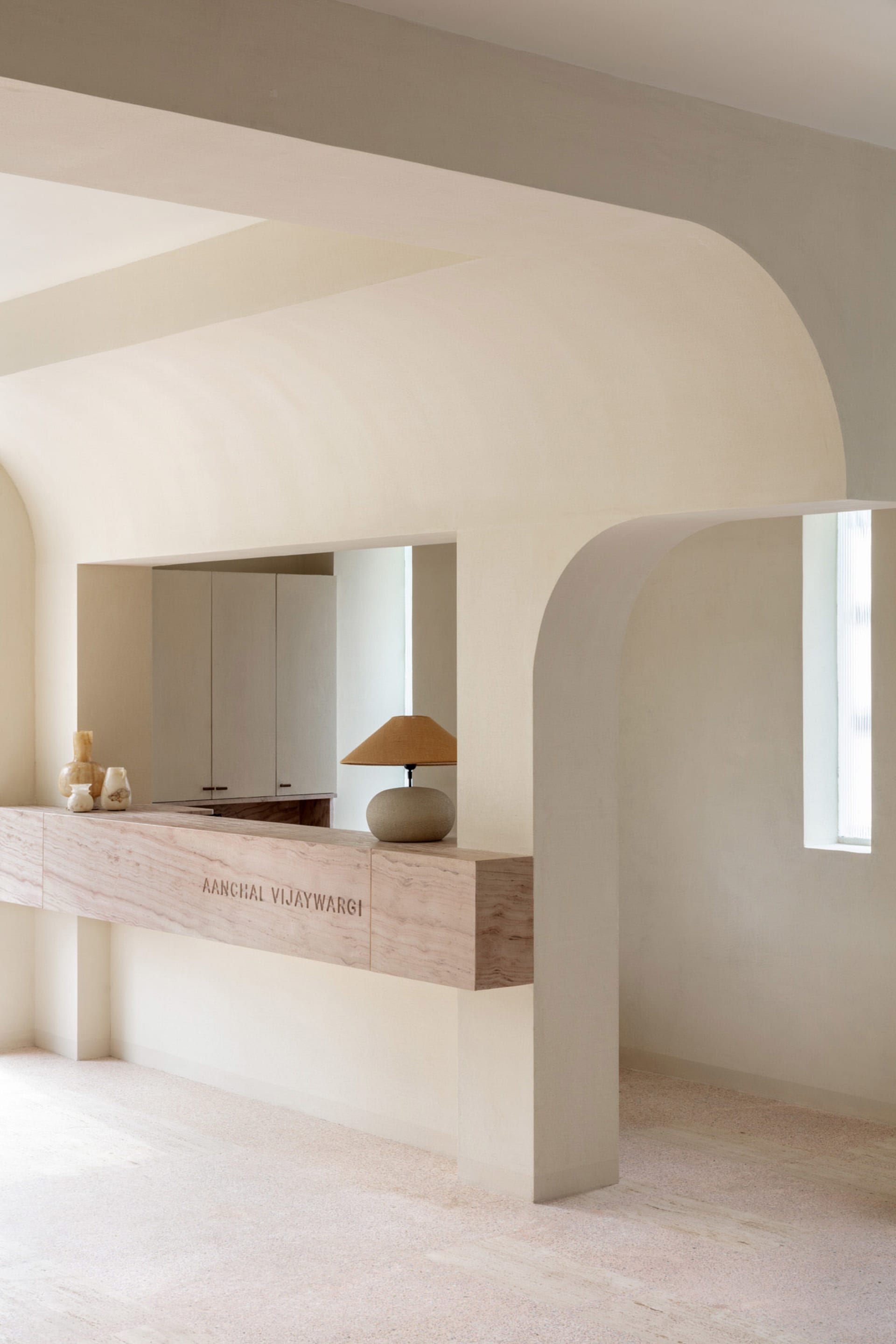 A minimalist interior space with neutral tones, featuring a wooden counter with a lamp and decorative items, and an arched do