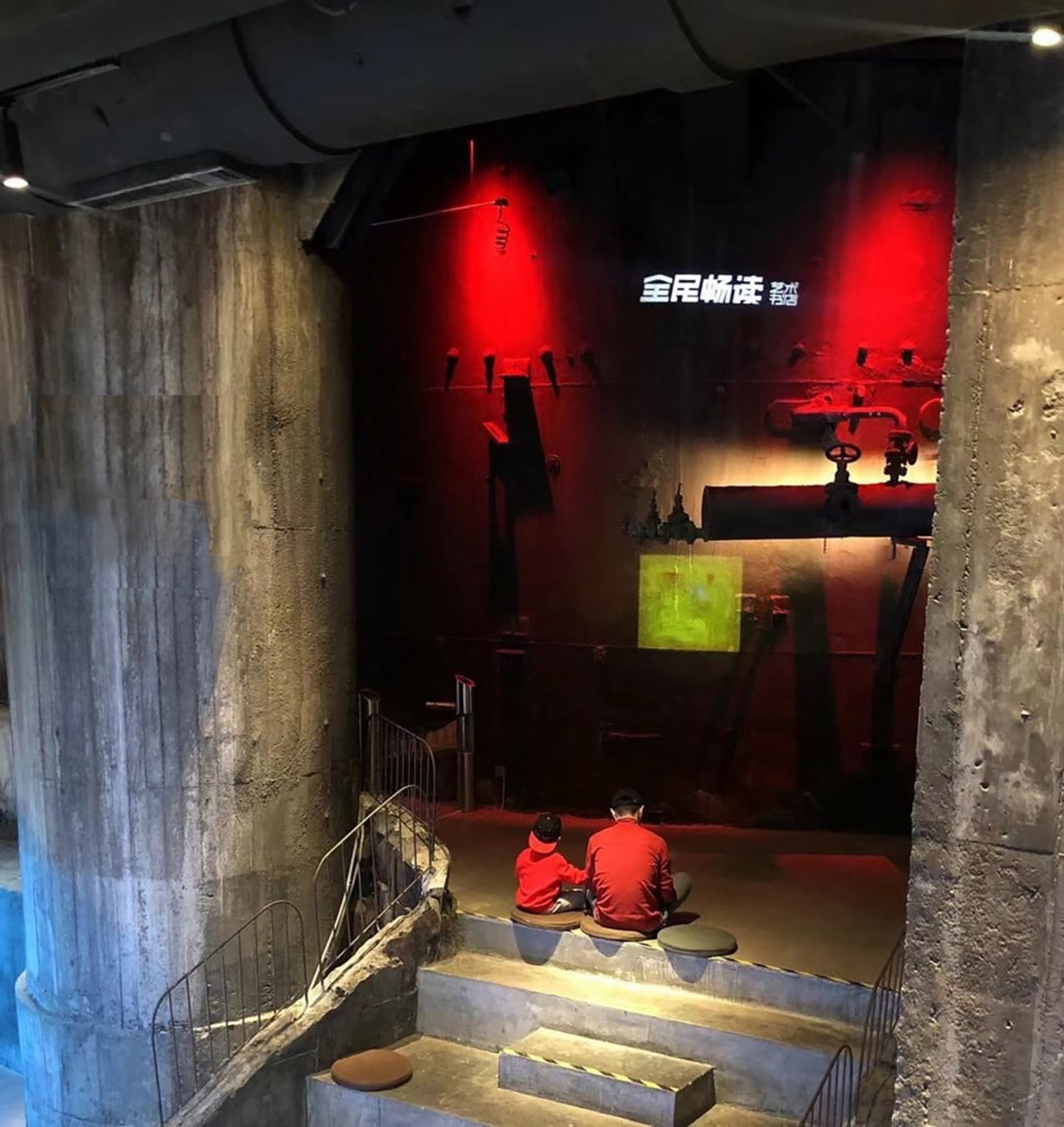 兩位人士坐在博物館展覽中的階梯上，展品包括混凝土管與紅光照亮的畫作。
