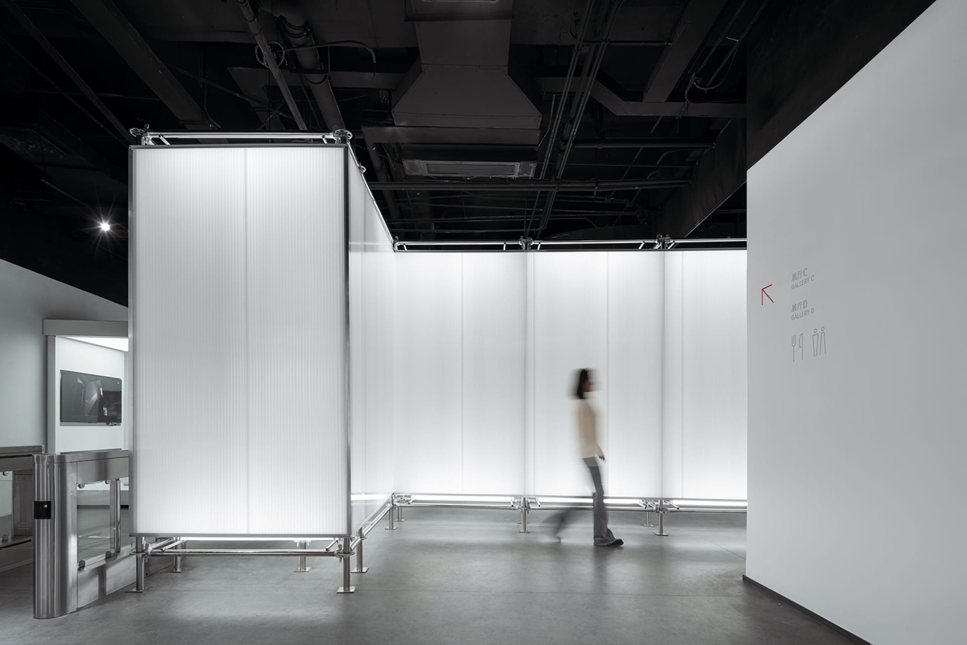 A minimalist exhibition space featuring glass walls and a single blurred person walking through the space.
