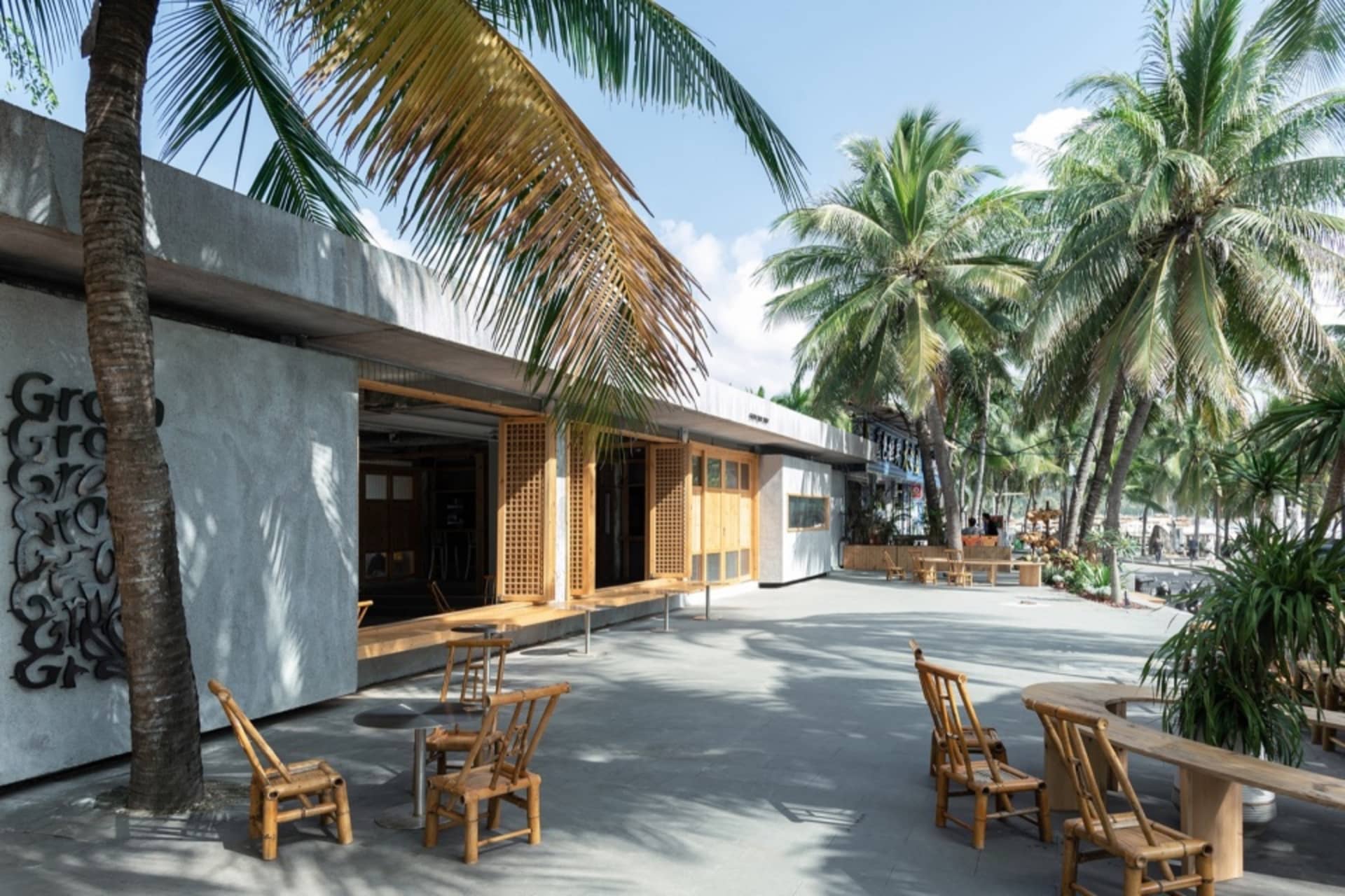 Outdoor seating area of a minimalist street-side café with concrete walls, bamboo chairs, and palm trees in the background.