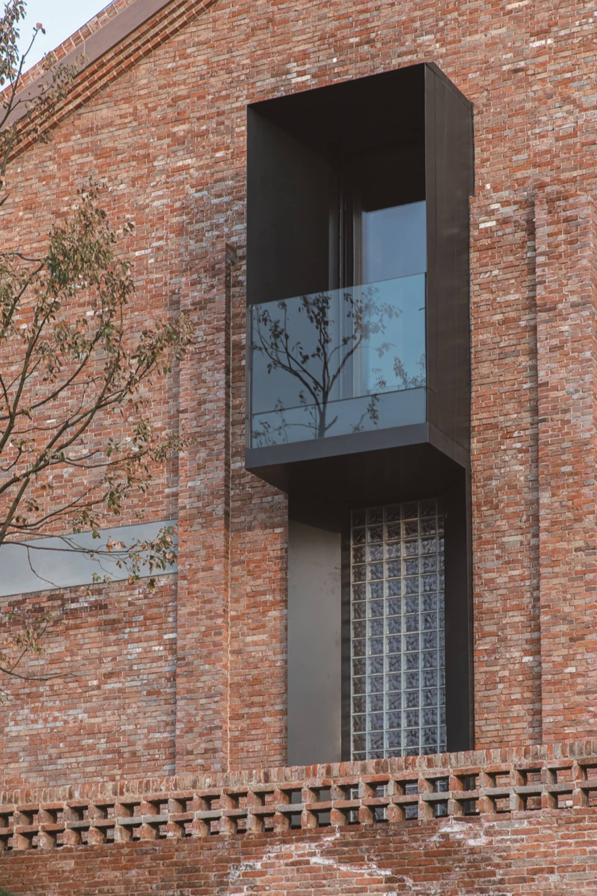 A modern building with a facade made of red bricks. A glass window with black frame and a glass balcony are visible.