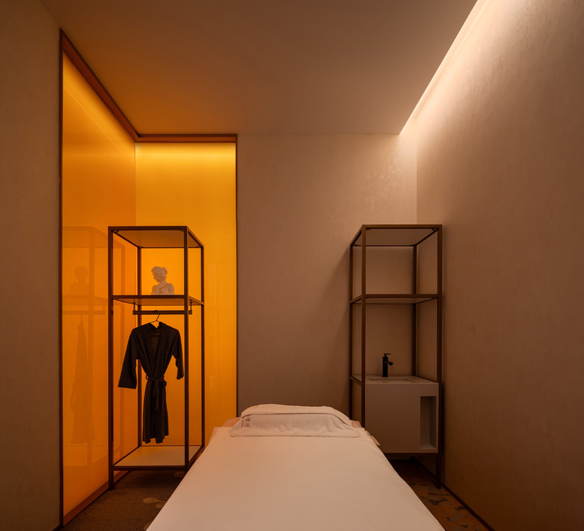 A minimalist spa treatment room with wooden furniture and white walls, featuring a massage table and a glass-enclosed closet.