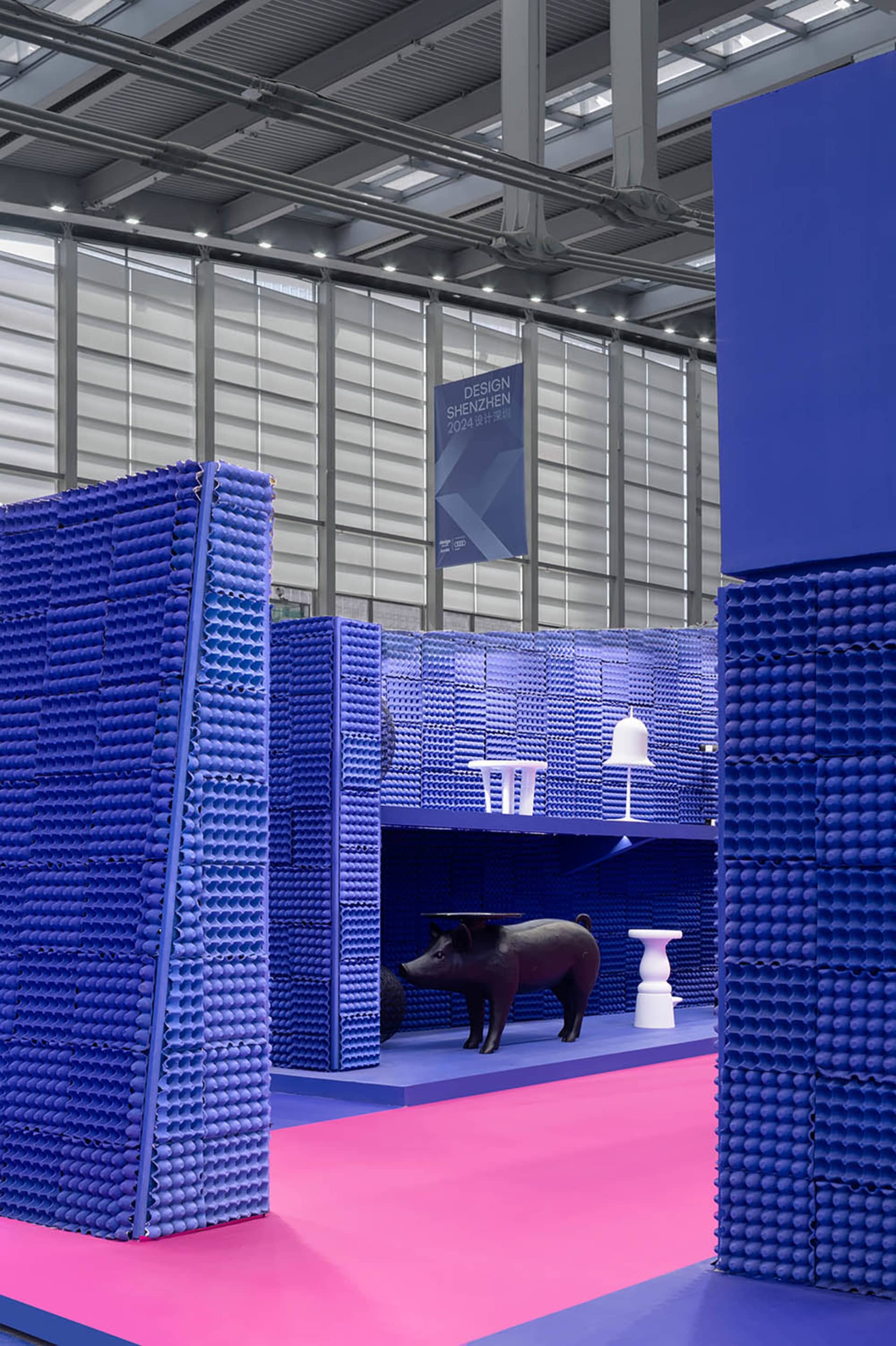 A minimalist installation featuring blue plastic structures and a black pig sculpture in a large space with pink flooring.