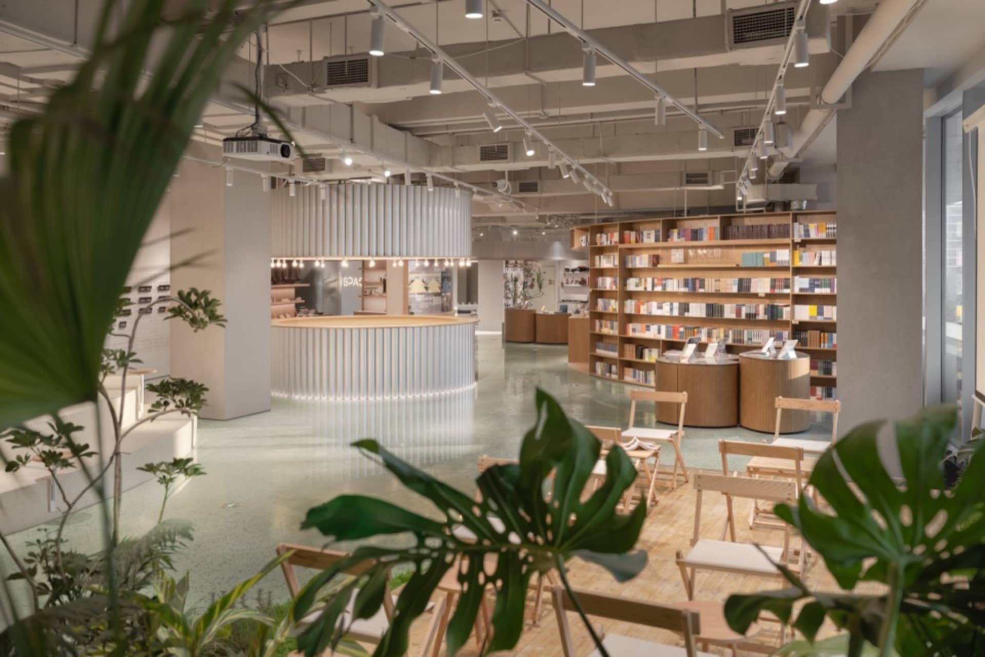 A modern cafe lounge with concrete flooring and wooden furniture, featuring green plants and a central circular bar.