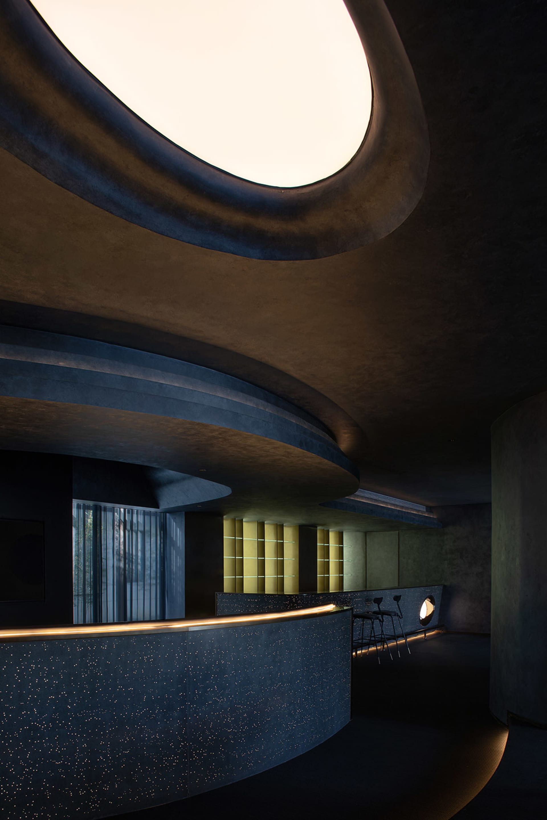Modern hotel lobby interior featuring a sleek, curved concrete reception desk with a skylight overhead.