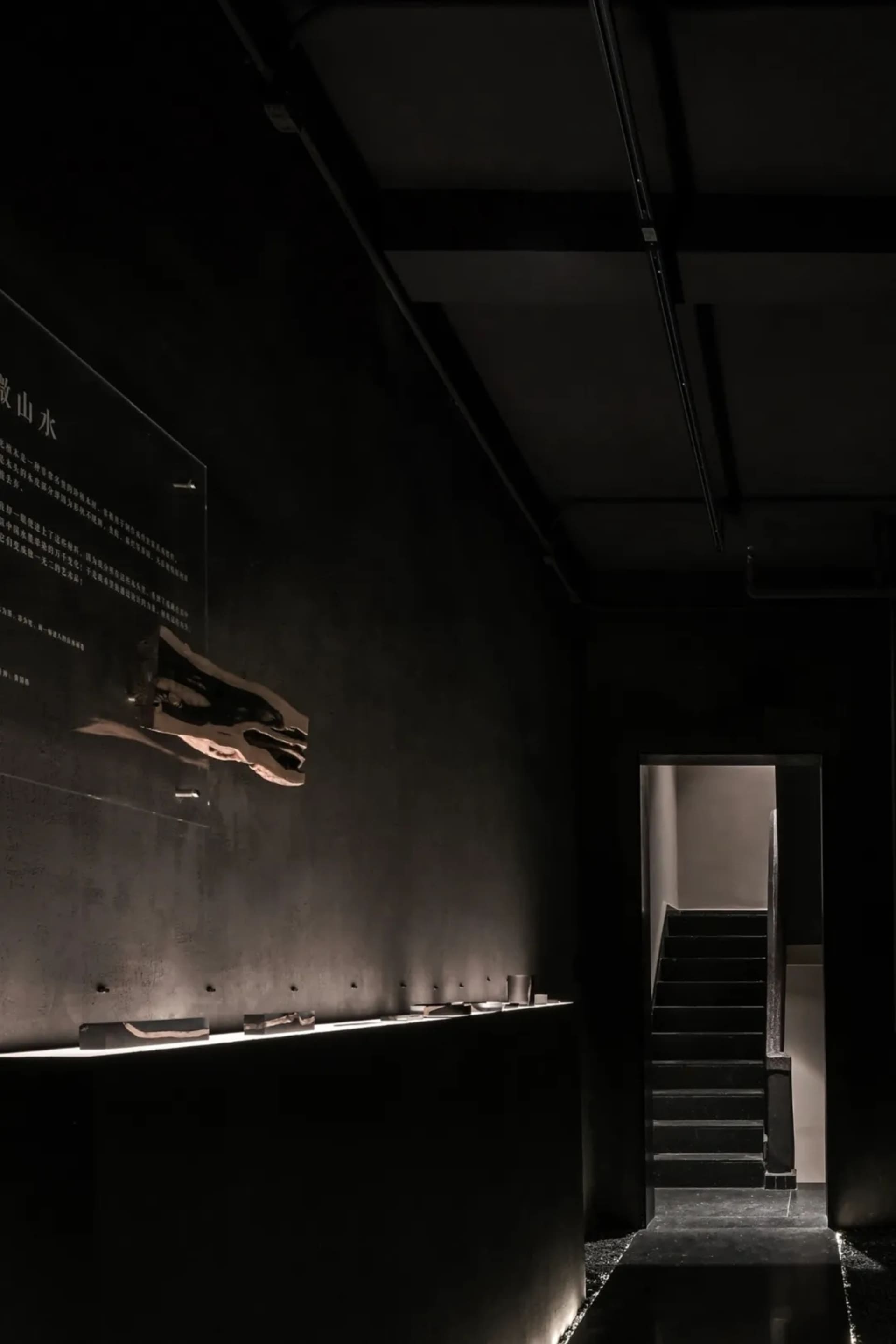 A minimalistic architecture gallery with a concrete wall and floor, featuring a display case with a wooden sculpture suspende