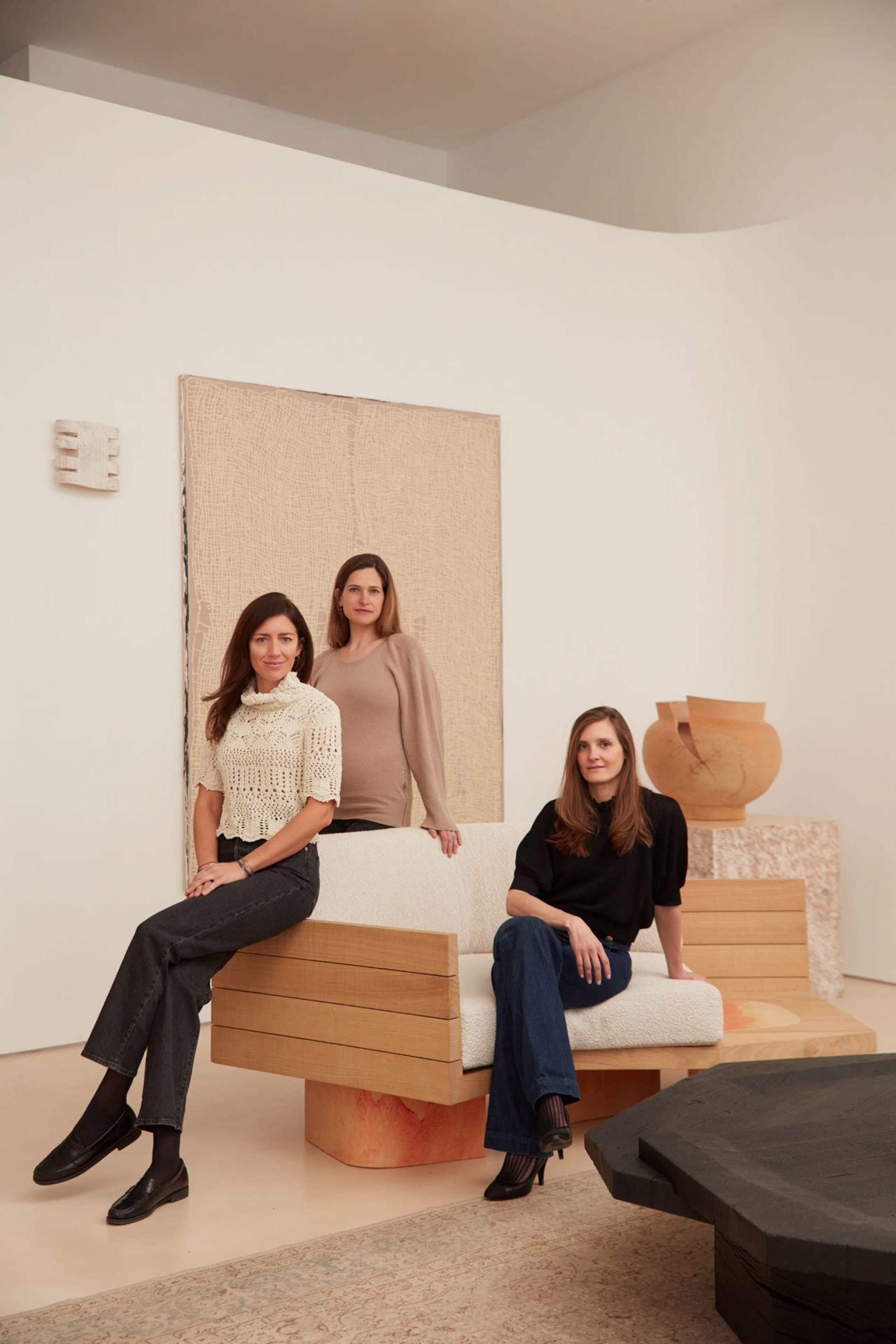 Three women posing in a contemporary interior setting with a wooden sofa and a textured wall hanging.